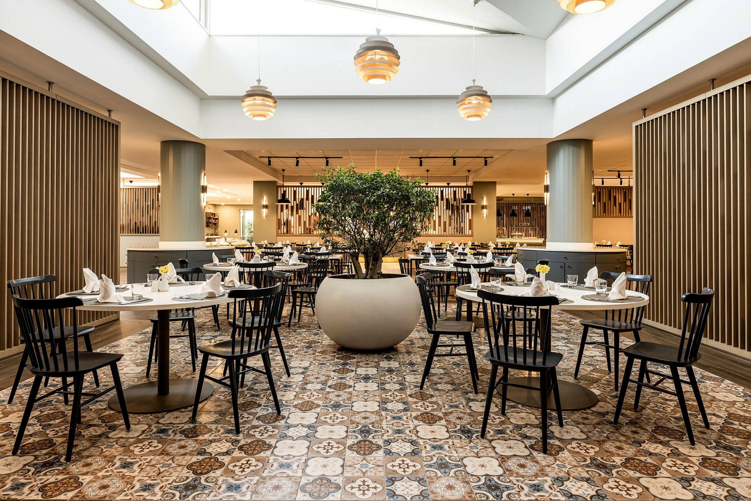 Billede af en restaurant med dækkede borde og en stor potte med grønne planter i midten. (AI-genereret tekst)