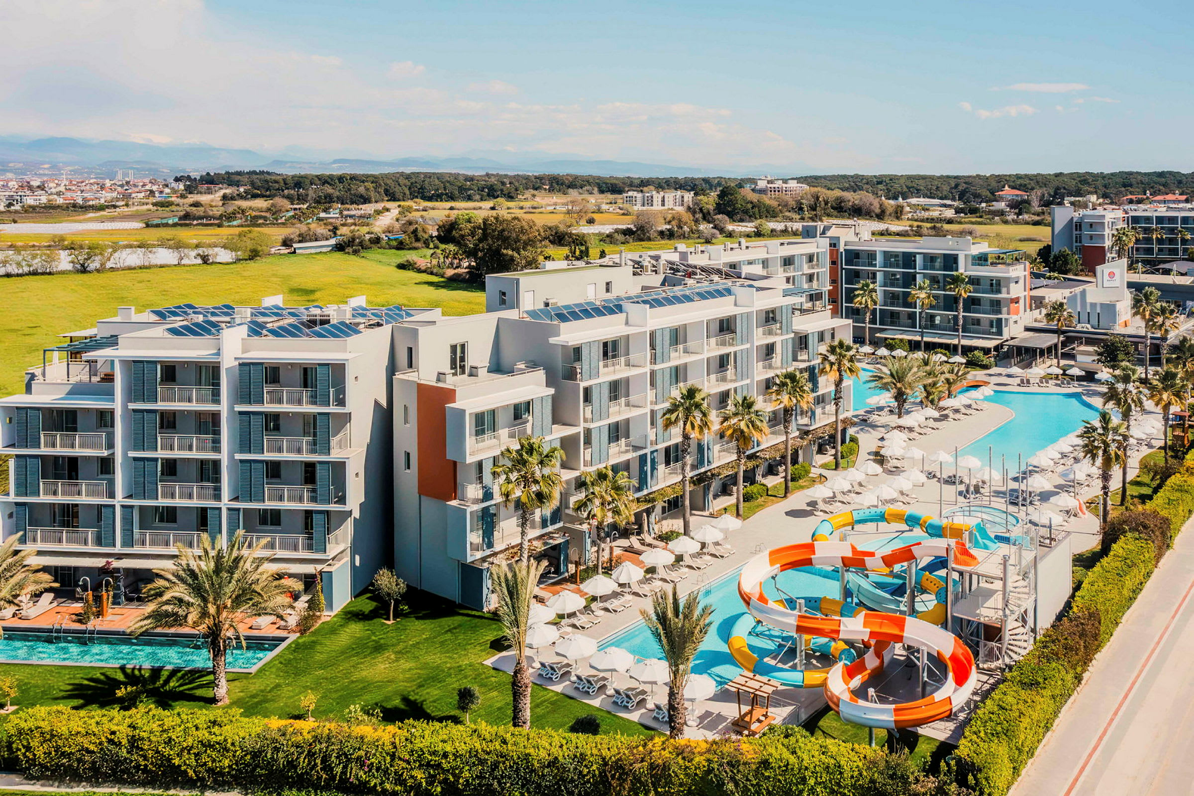 Luftfoto af et hotel med pools og vandrutsjebaner, omgivet af grønne områder. (AI-genereret tekst)