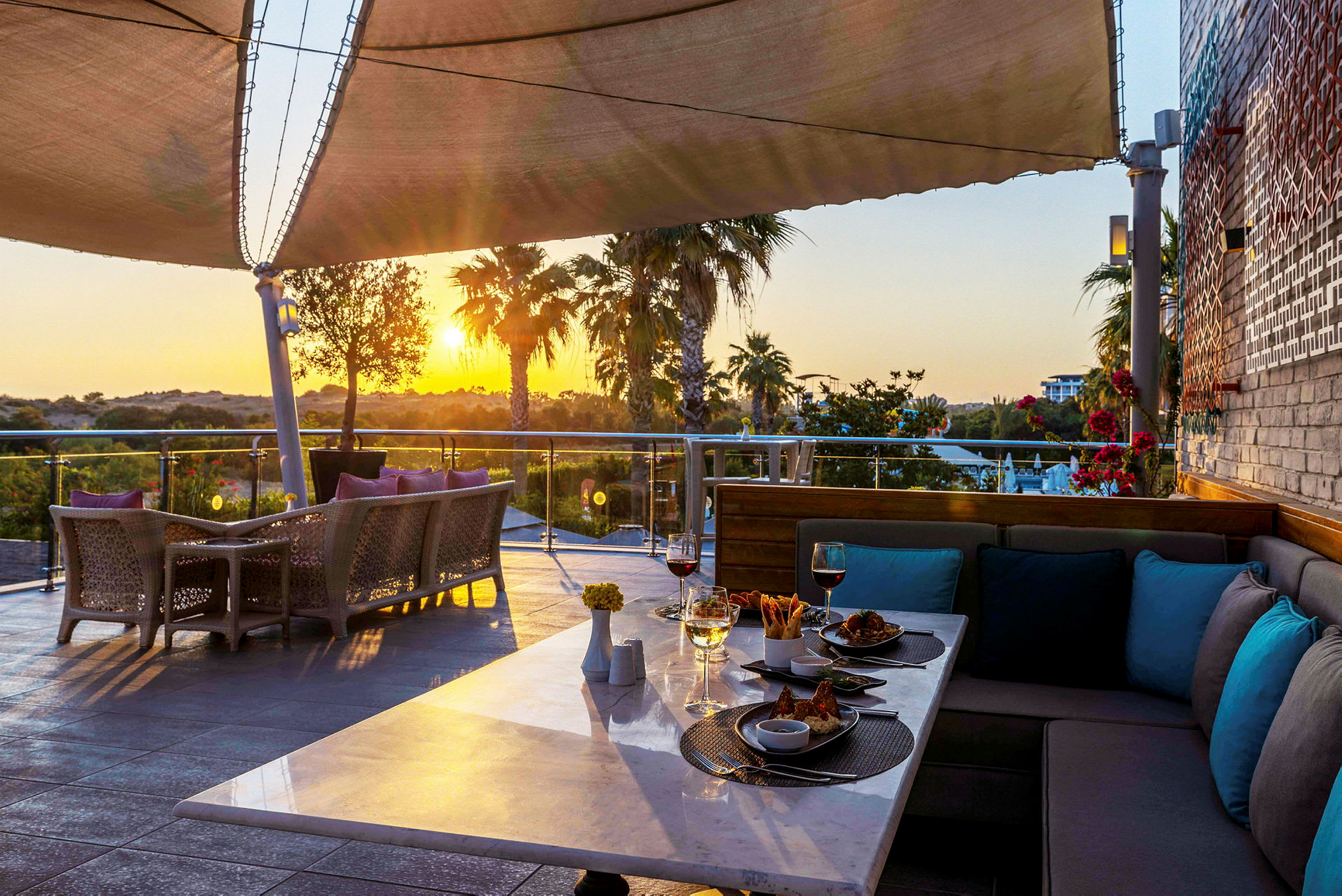 En terrasse med bord og stole, mad og drikke på et bord, med udsigt over en have og en solnedgang. (AI-genereret tekst)