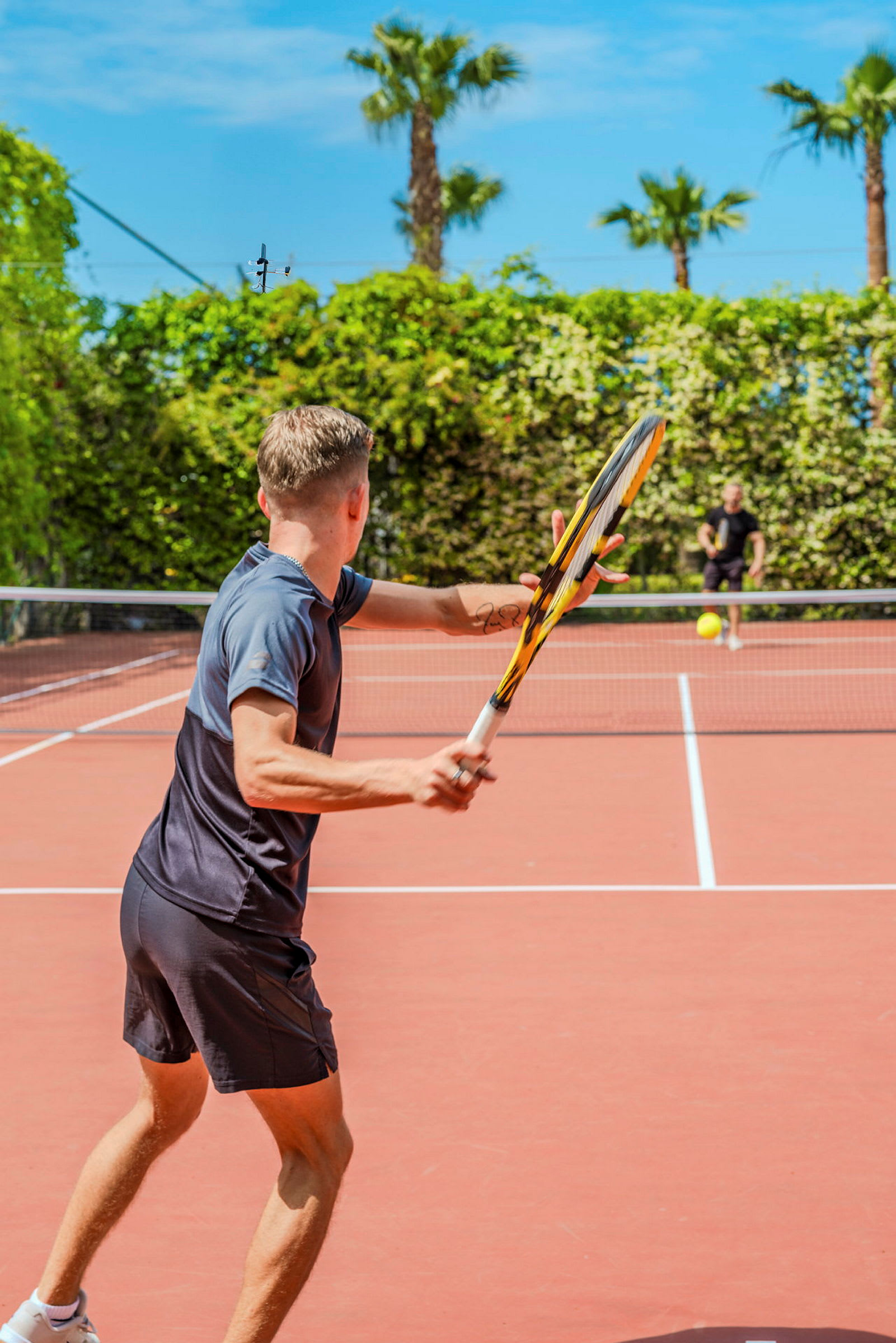 To personer spiller tennis udendørs på en tennisbane omgivet af beplantning og palmer. (AI-genereret tekst)