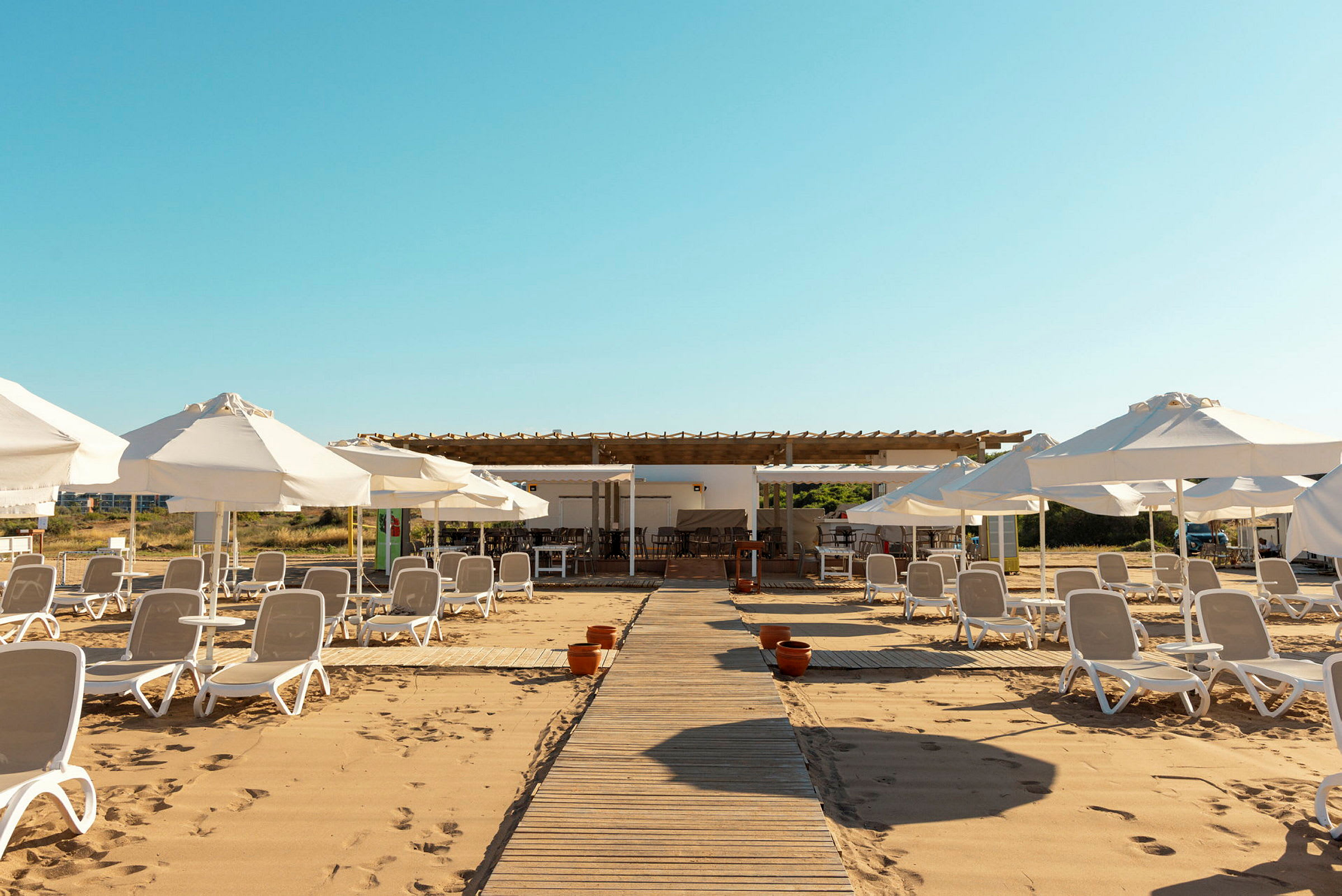 En strand med liggestole og parasoller på sanden samt en strandbar i baggrunden. (AI-genereret tekst)