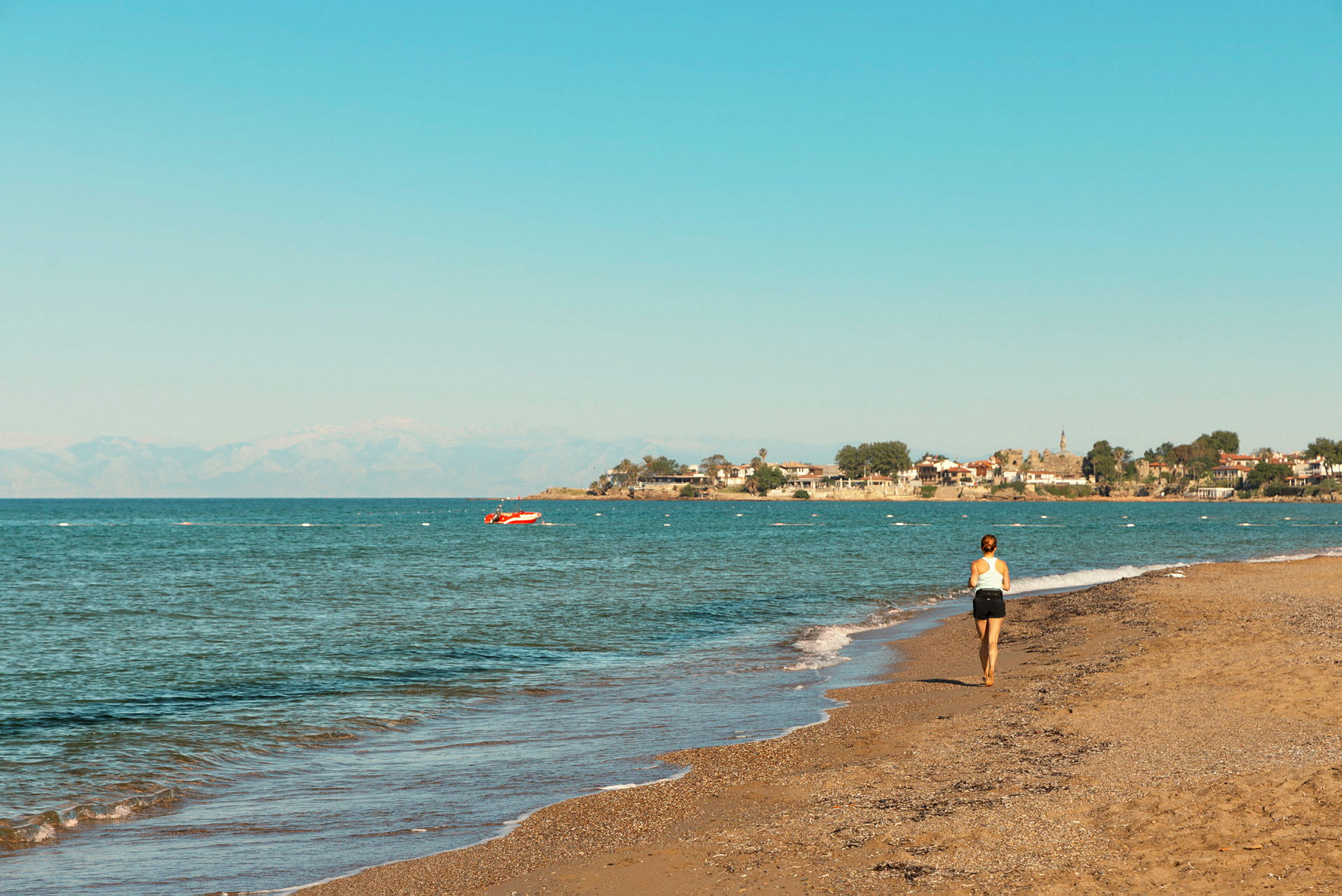 En kvinde går langs en strand med udsigt over havet og en landsby ved kysten i baggrunden. (AI-genereret tekst)