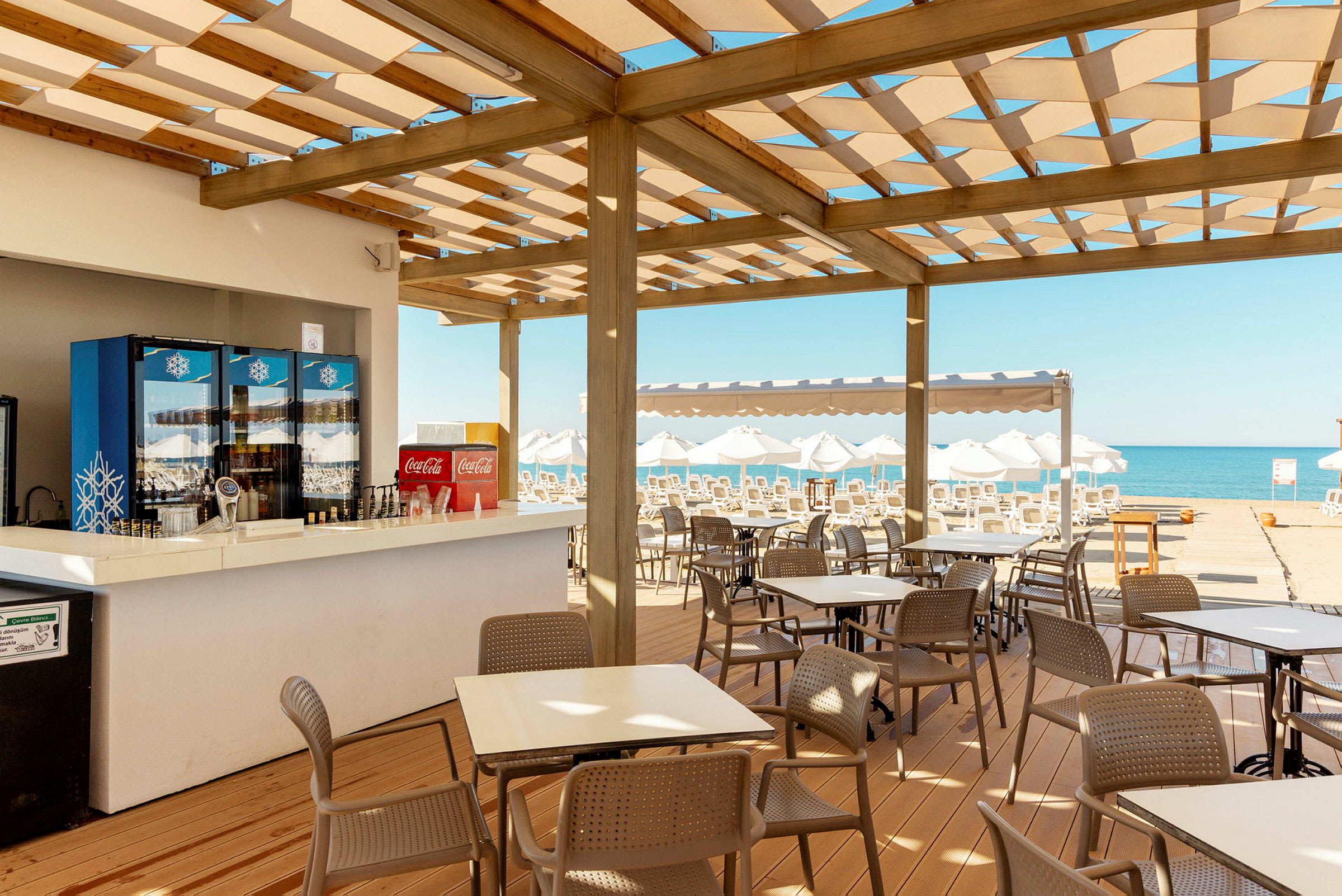 En café med udendørs servering under et trætagesystem nær stranden. Bord og stole er opstillet, og i baggrunden ses liggestole og havet. (AI-genereret tekst)