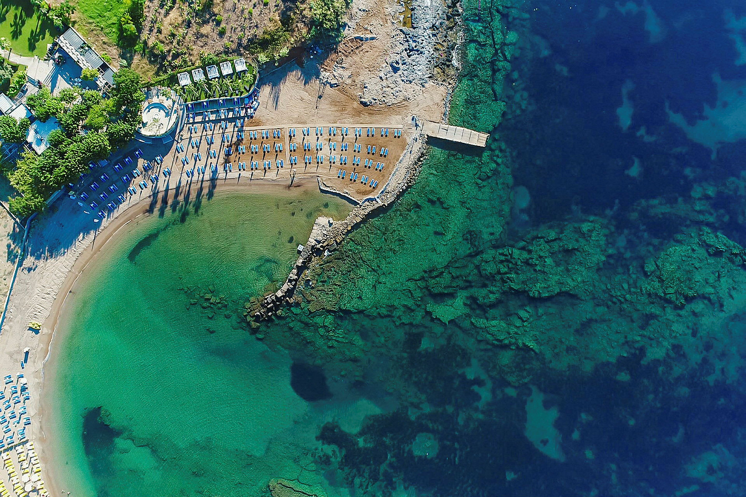 Luftfoto af en strand med liggestole og parasoller ved en turkisblå hav. (AI-genereret tekst)