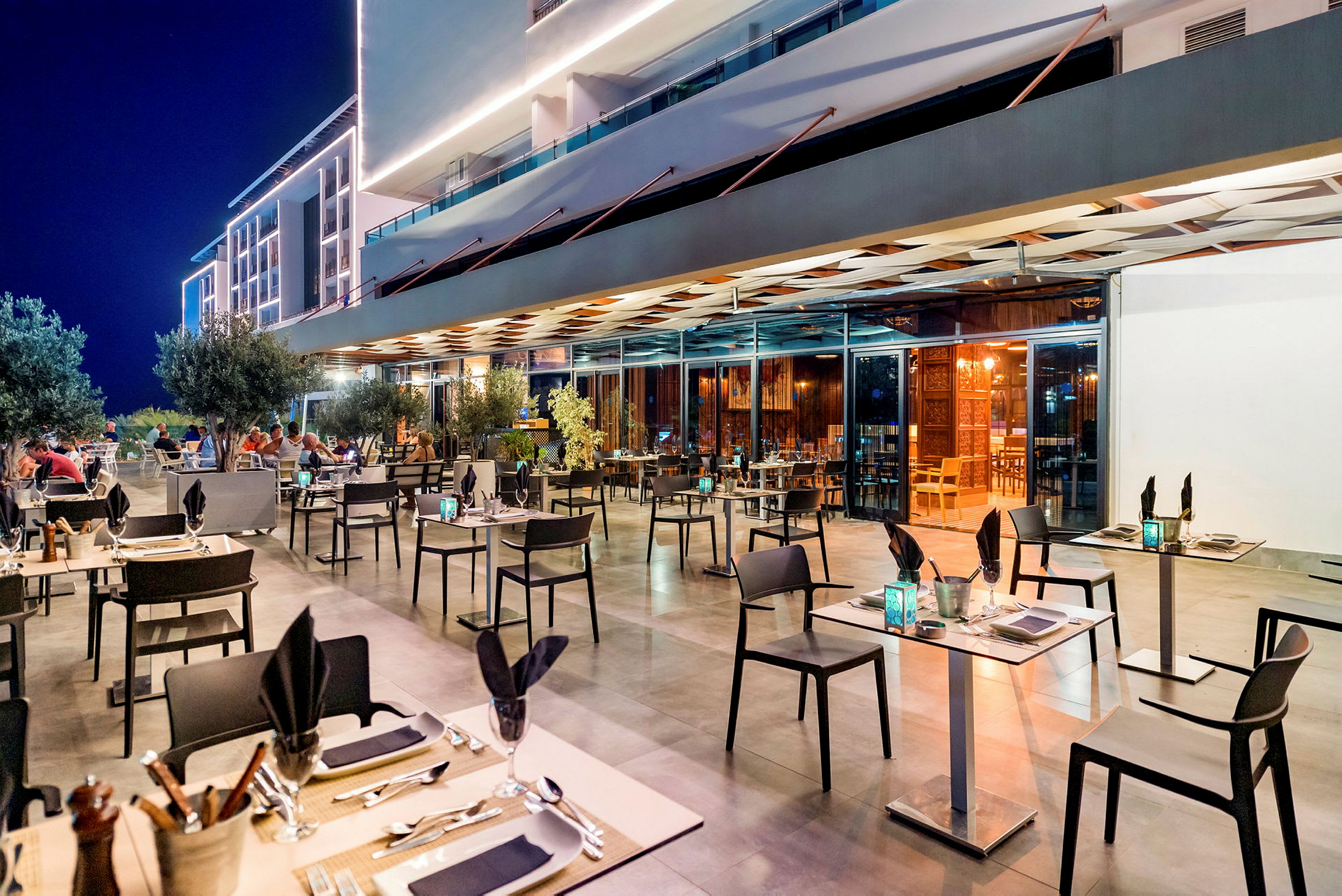 Udendørsrestaurant med oplyst facade og borde opstillede på terrassen. (AI-genereret tekst)