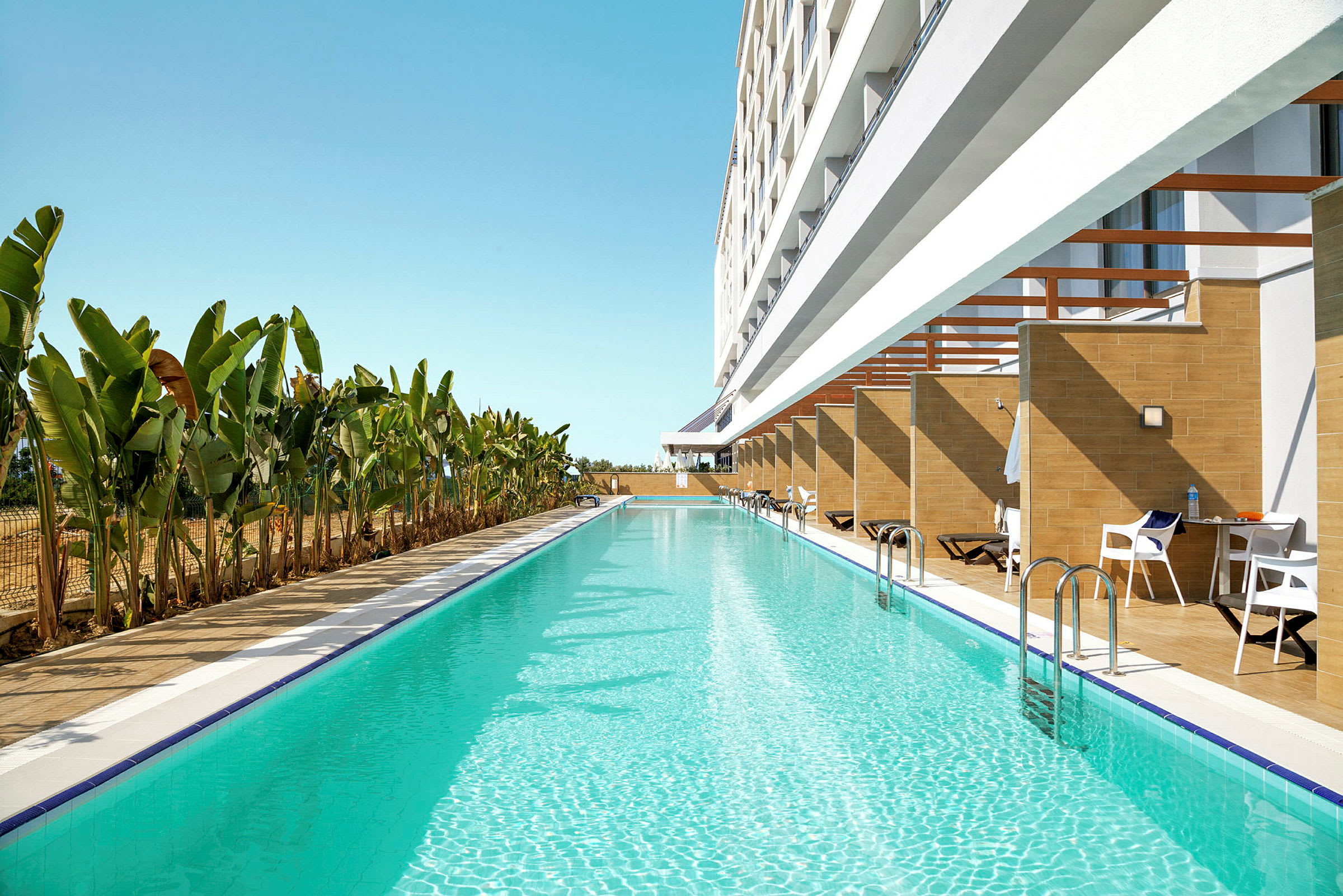 Lang swimmingpool med klart blåt vand ved siden af en bygning med private terrasser og liggestole, omgivet af banantræer. (AI-genereret tekst)