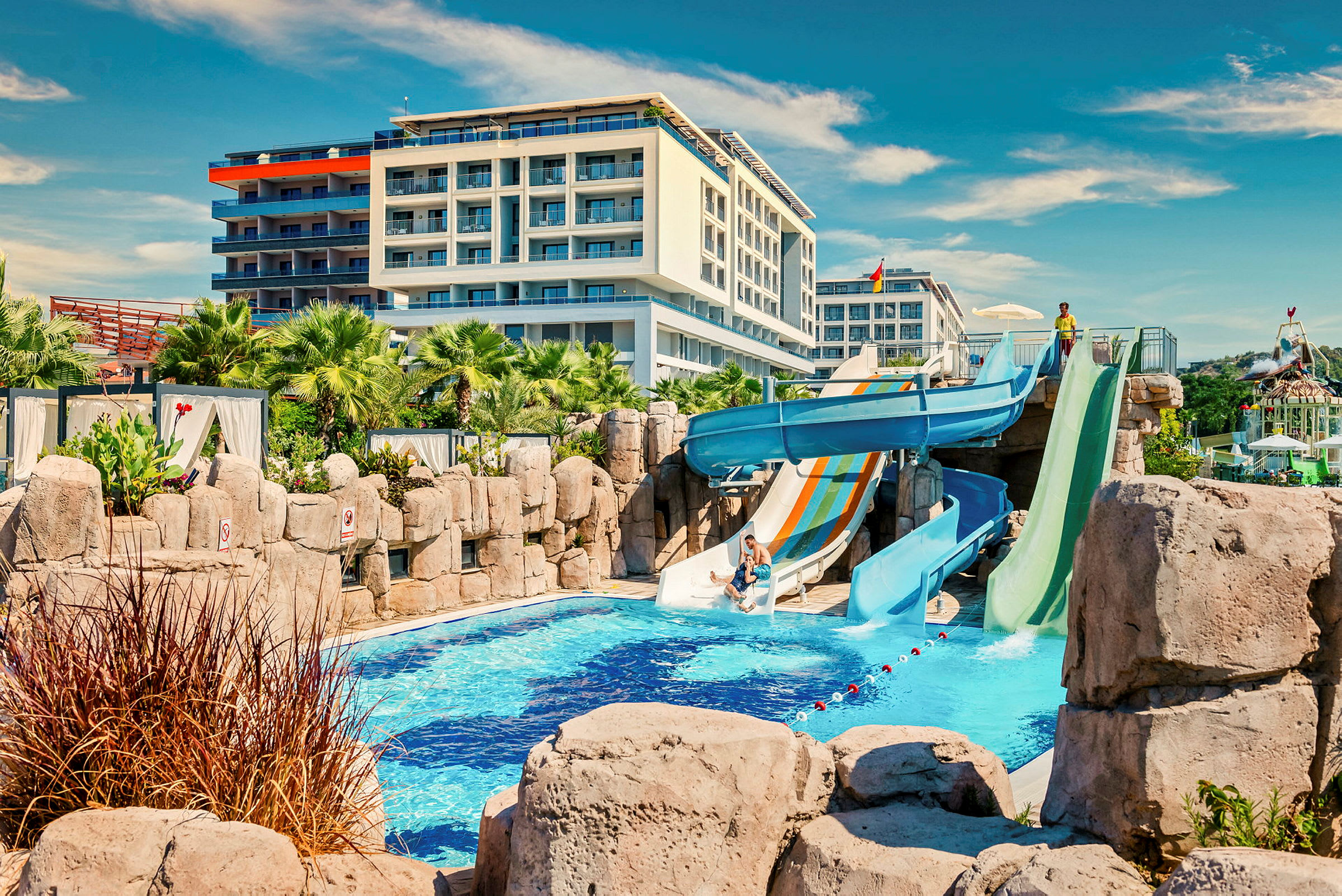 Hotel med poolområde med vandrutsjebaner, grønne palmer og bygning i baggrunden. (AI-genereret tekst)