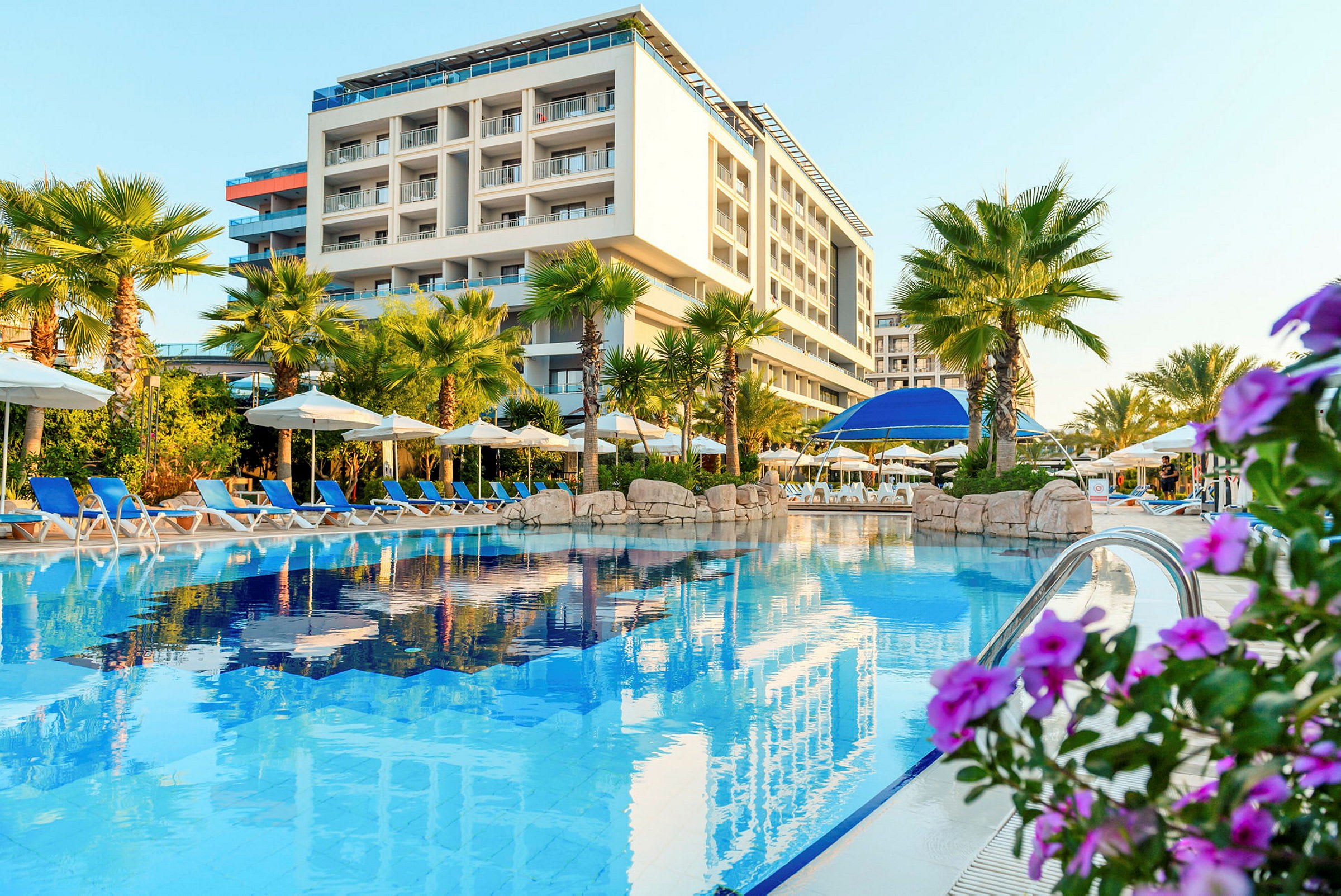 Billede af en hotelbygning bag en pool med liggestole og parasoller samt omkringliggende palmer og blomster. (AI-genereret tekst)
