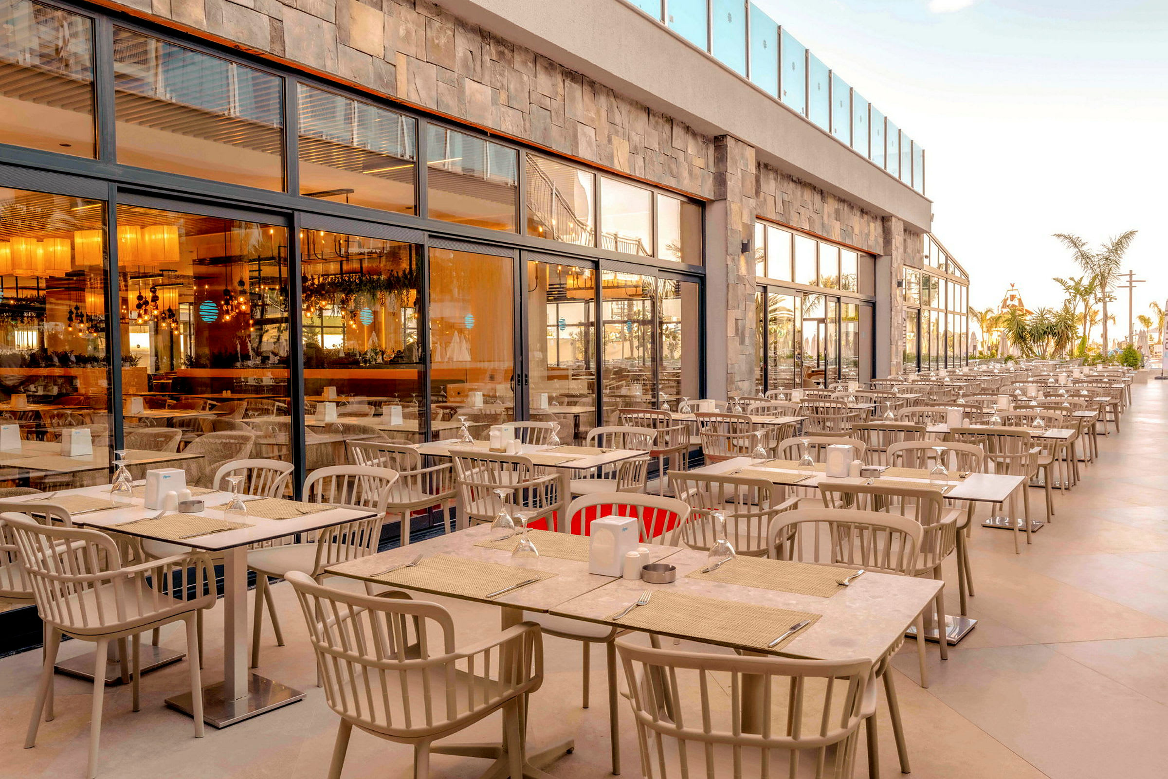 En udendørs terrasse med mange borde og stole foran glasfacaden af en restaurant. (AI-genereret tekst)