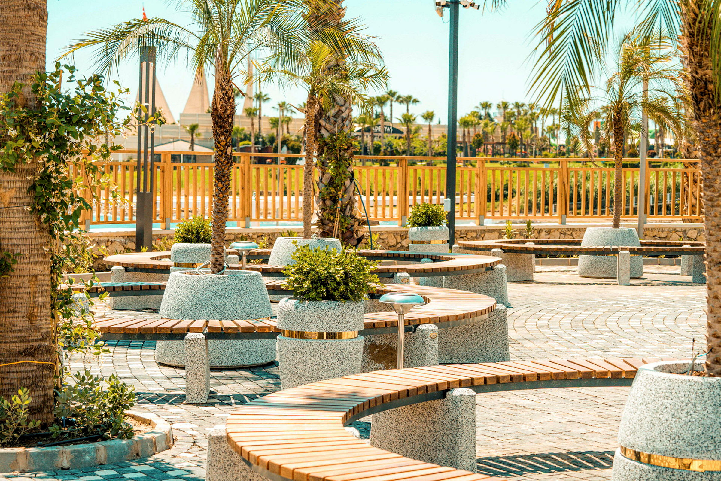 Parkområde med runde træbænke og beplantning omgivet af palmer. (AI-genereret tekst)