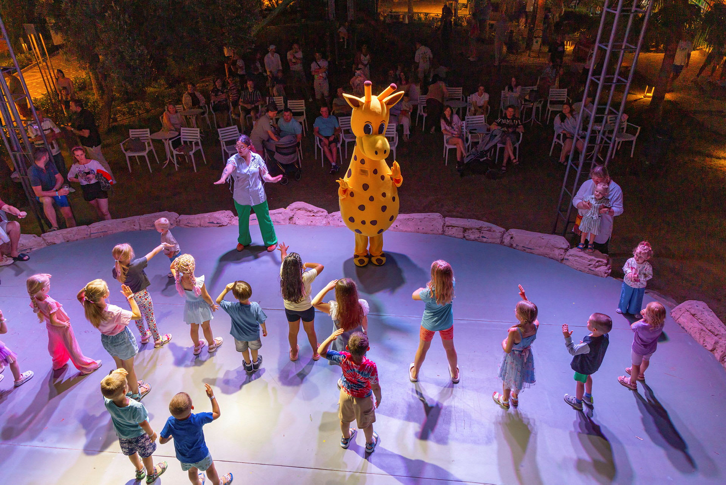 En gruppe børn danser omkring en maskot, der forestiller en gul giraf, på en scene, mens voksne sidder og kigger på i baggrunden. (AI-genereret tekst)