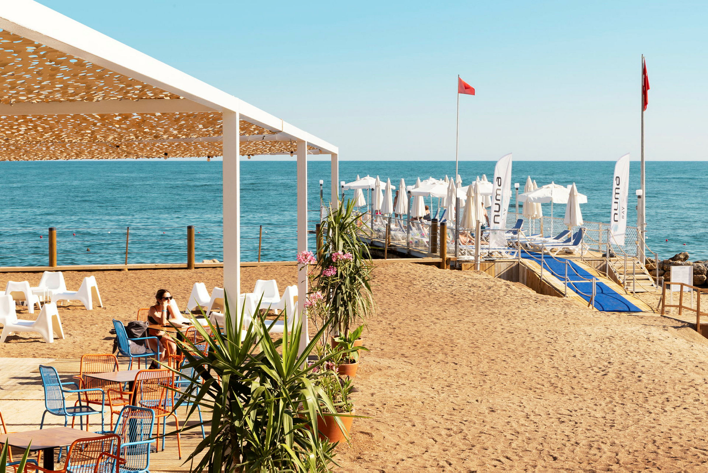 En strand med stole og borde under en pergola, og i baggrunden ses liggestole og parasoller ved havet. (AI-genereret tekst)