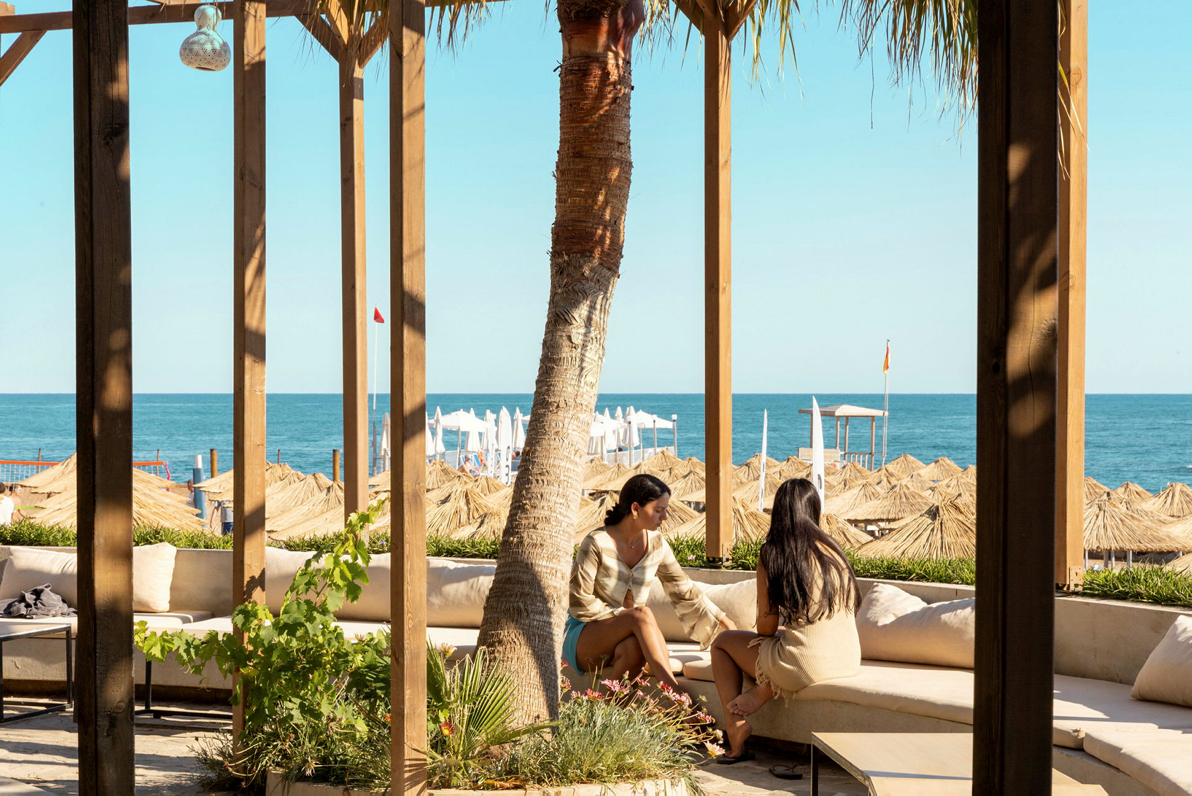 To personer sidder på en udendørs sofa under en træpergola og ser ud mod havet og stranden med parasoller. (AI-genereret tekst)