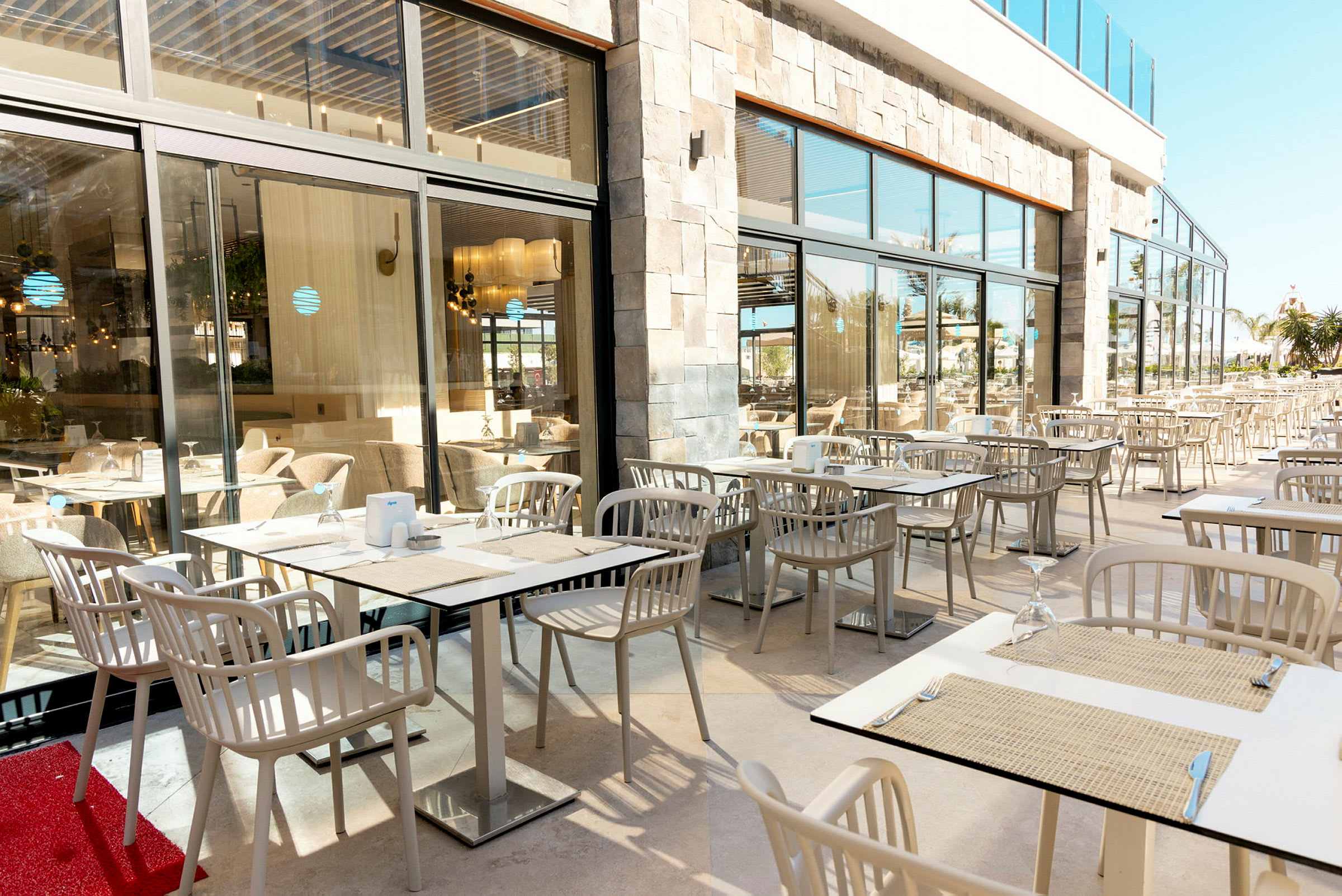 En udendørsterrasse med borde og stole ved siden af et stort vindue, der viser restaurantens indendørsområde. (AI-genereret tekst)