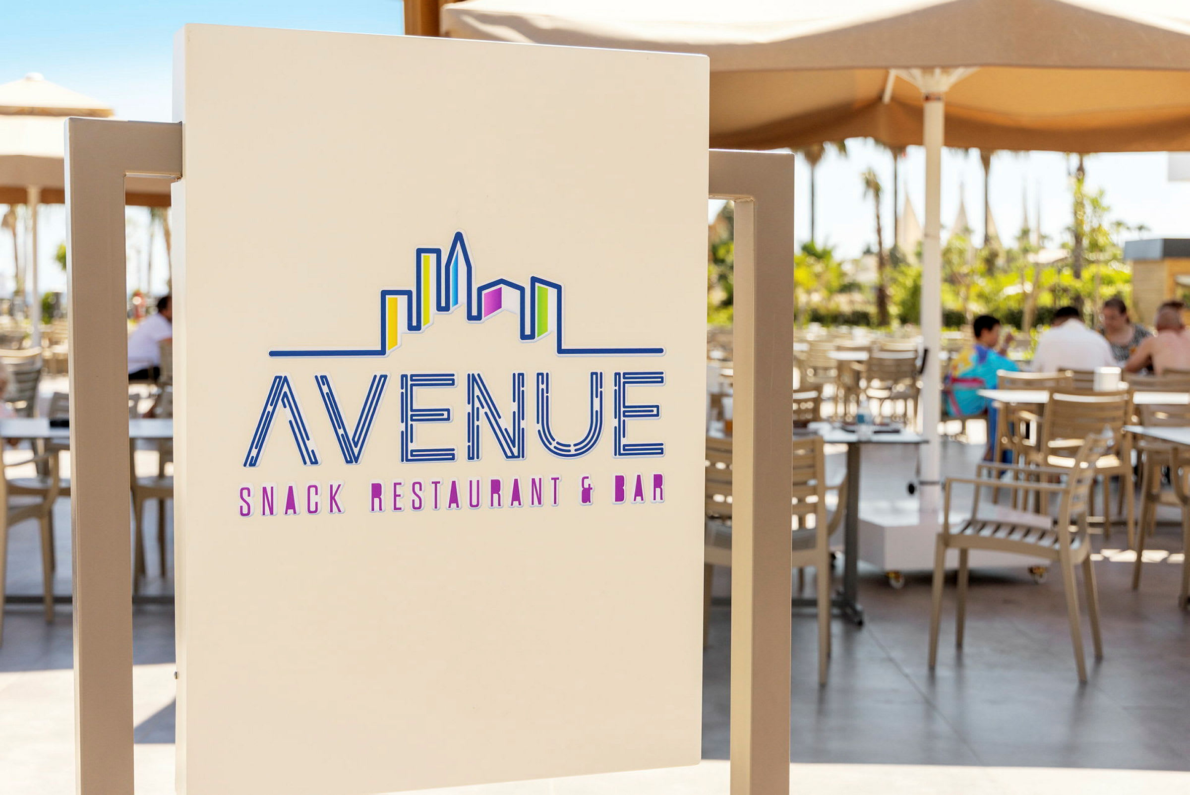 Et skilt, der viser teksten "Avenue Snack Restaurant & Bar" med en stiliseret skyline-motiv. I baggrunden ses en udendørsservering med borde og stole under parasoller. (AI-genereret tekst)
