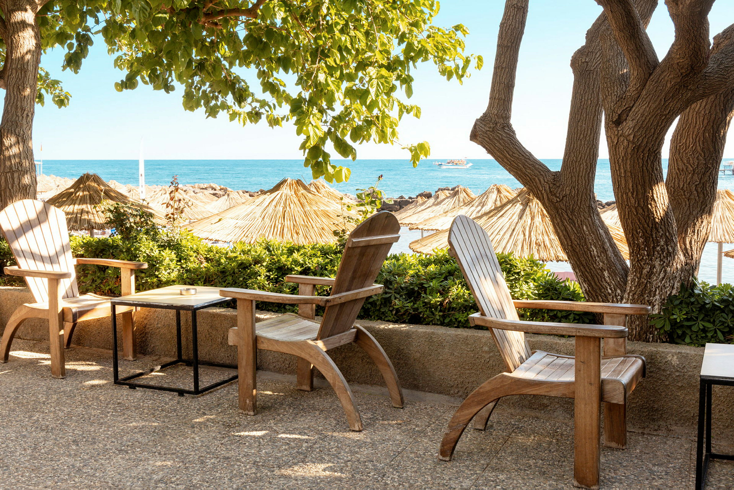 Træstole og borde står på en terrasse med udsigt over en strand og havet, omgivet af træer og parasoller. (AI-genereret tekst)