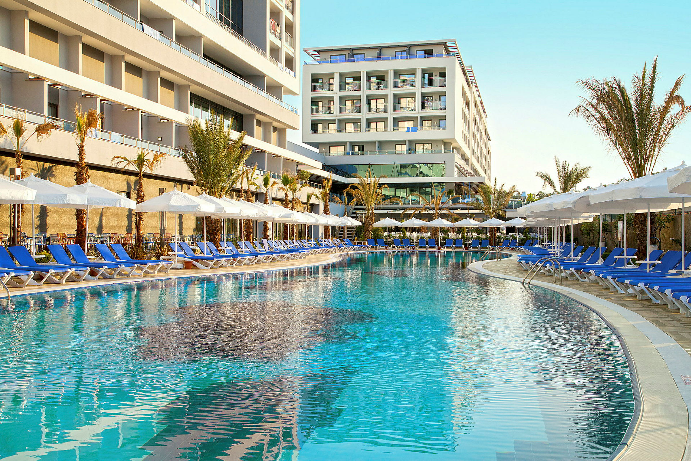 Udendørs pool med liggestole omkring samt hotelbygninger i baggrunden. (AI-genereret tekst)