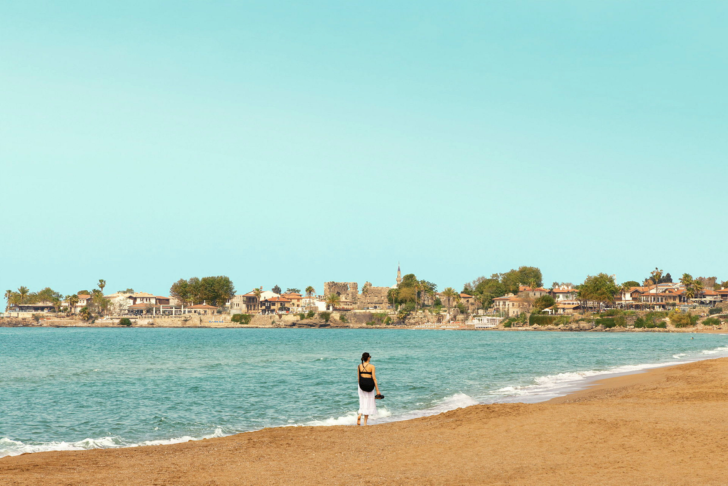 Person står på stranden ved havet med bygninger i baggrunden. (AI-genereret tekst)