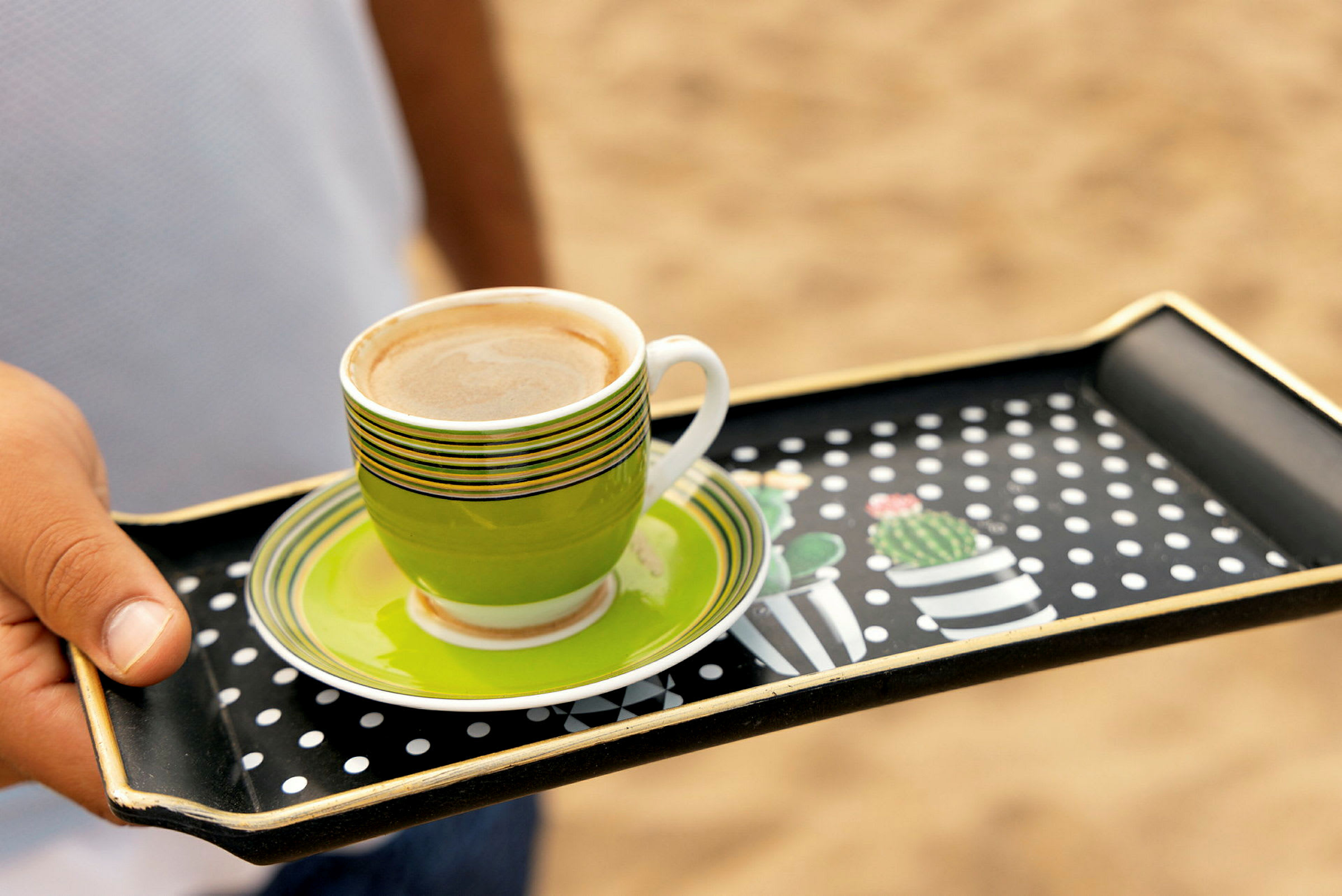 En person holder en bakke med en grøn kaffekop på en sandstrand. (AI-genereret tekst)
