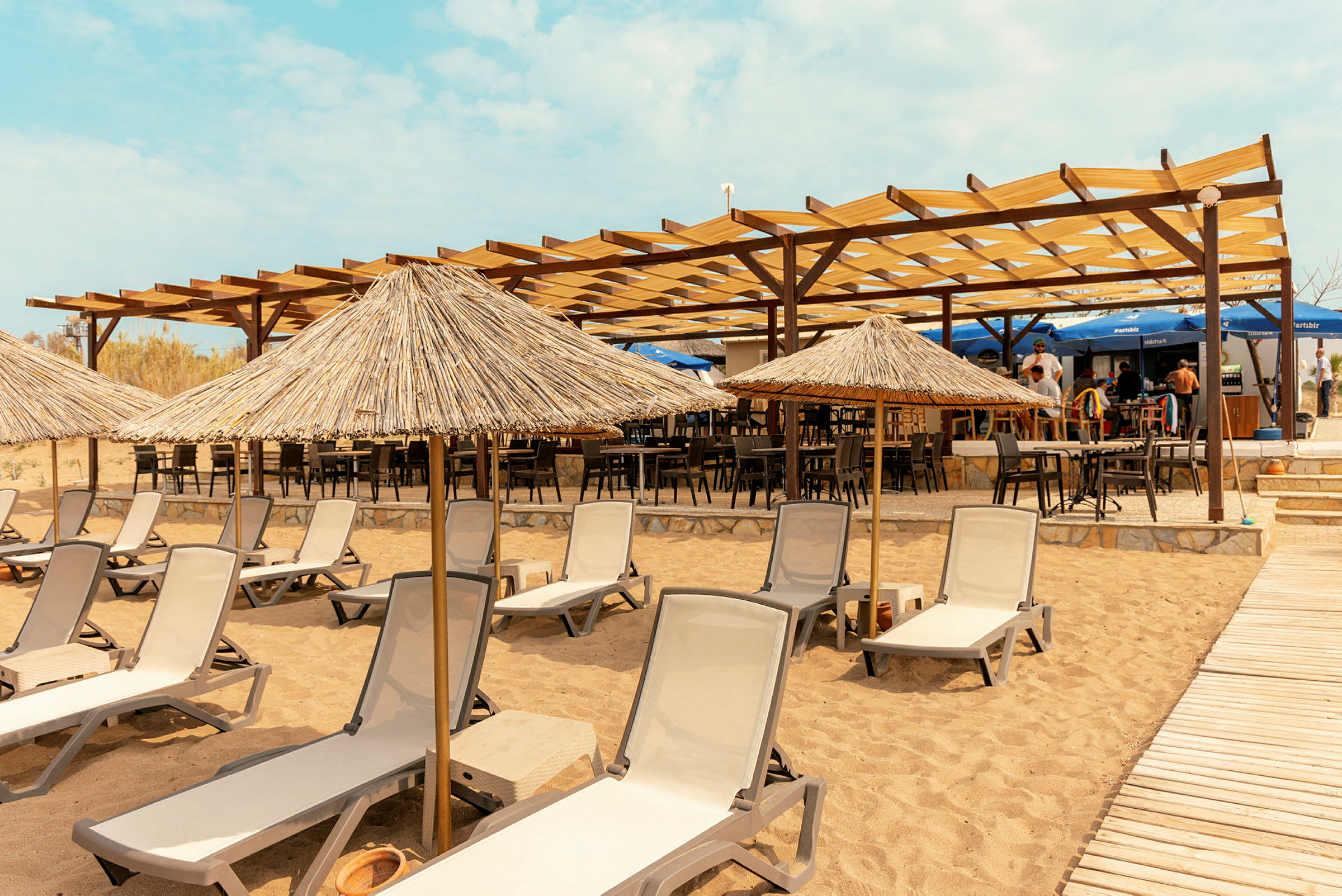 Liggestole og parasoller på en sandstrand med en café i baggrunden. (AI-genereret tekst)