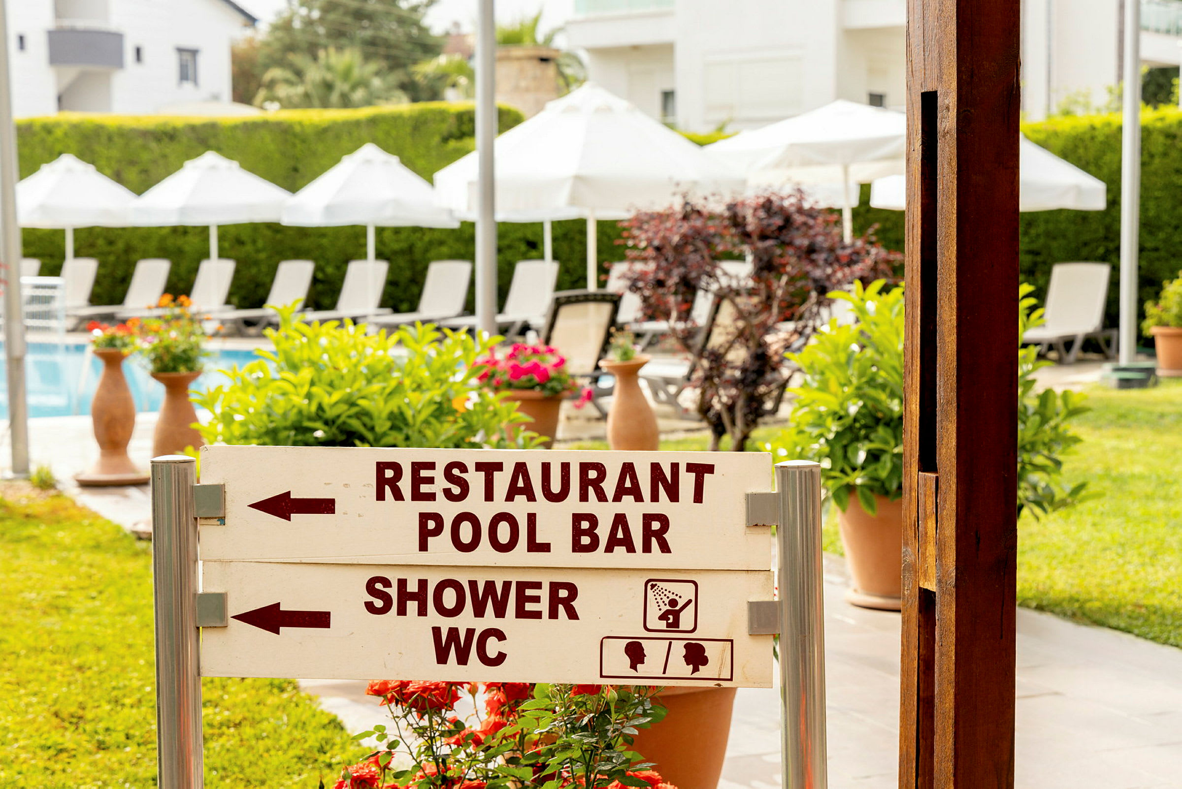 Skilt i en have med teksten 'Restaurant Pool Bar' og 'Shower WC', liggestole og parasoller ses i baggrunden. (AI-genereret tekst)