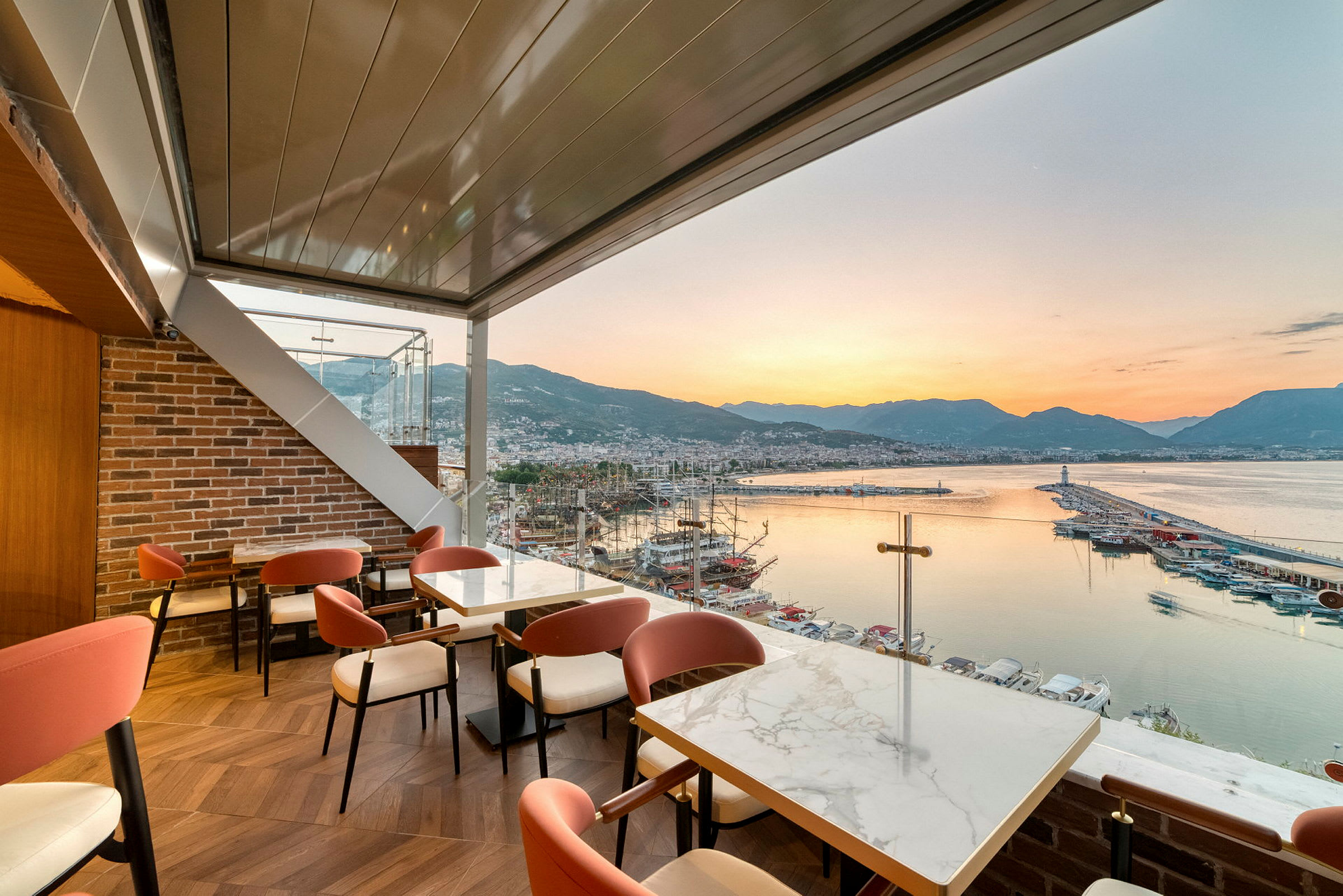 Restaurant med udsigt over en havn, havet og bjergene ved solnedgang. (AI-genereret tekst)