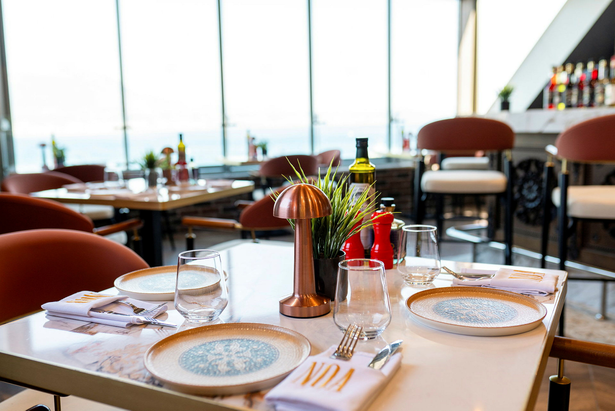 Bord på en restaurant med tallerkener, glas og bestik, udsigt mod store vinduer. (AI-genereret tekst)