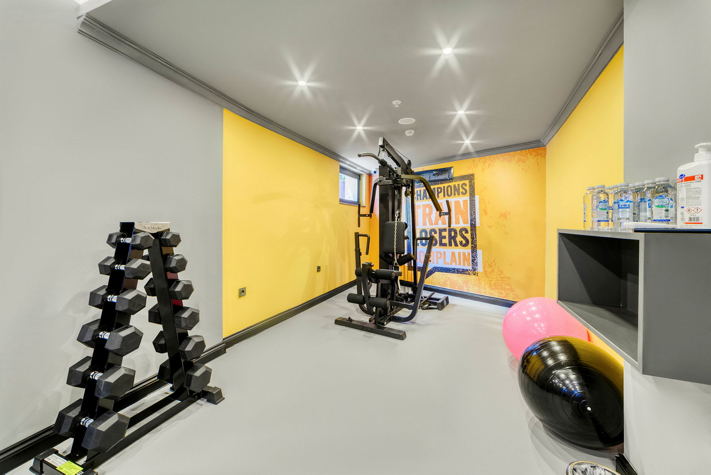 Et træningsrum med fitnessudstyr, håndvægte, en plakat på væggen og vandflasker. (AI-genereret tekst)