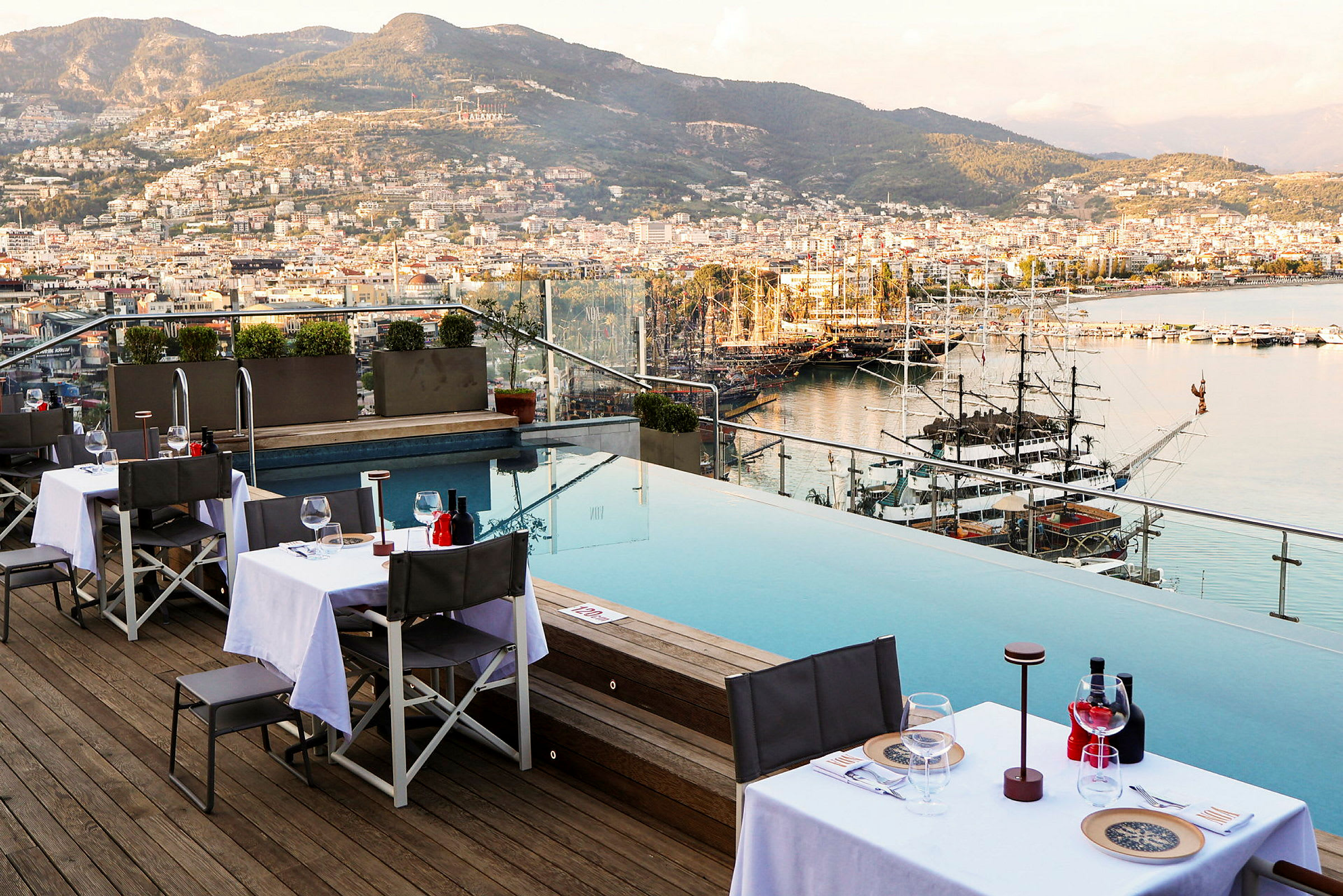 Udendørs spiseområde ved en infinitypool med udsigt over en by og havn. (AI-genereret tekst)