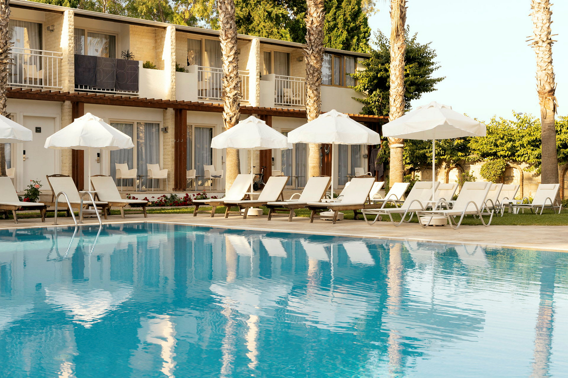 Billede af en hotelpool med liggestole og parasoller ved kanten, og bygninger i baggrunden. (AI-genereret tekst)