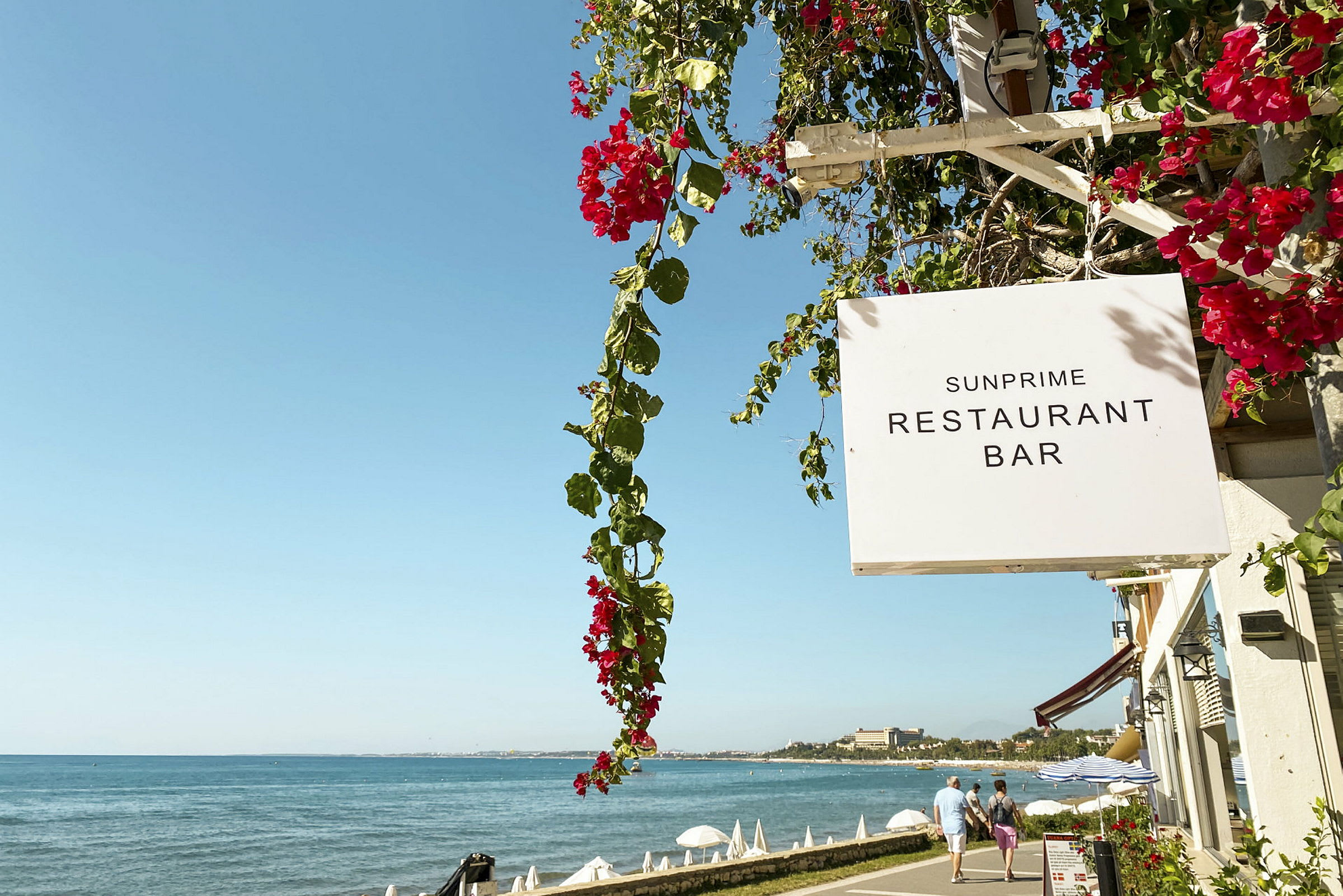 Skilt for Sunprime Restaurant Bar ved en promenade tæt på havet. (AI-genereret tekst)