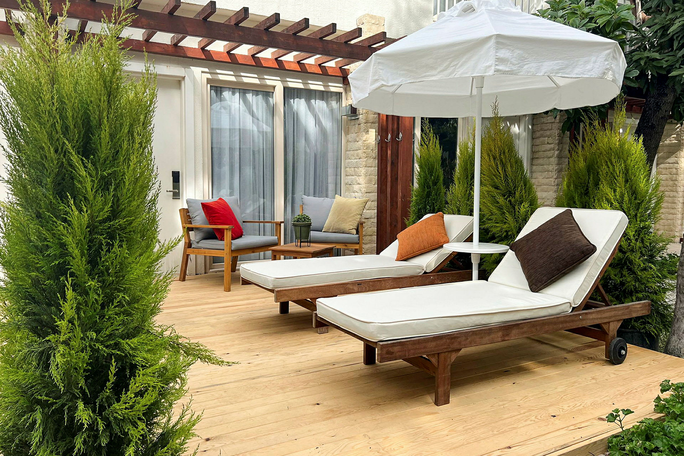 To liggestole med hvide hynder og puder står på en træterrasse under en parasol, omgivet af grønne planter. (AI-genereret tekst)