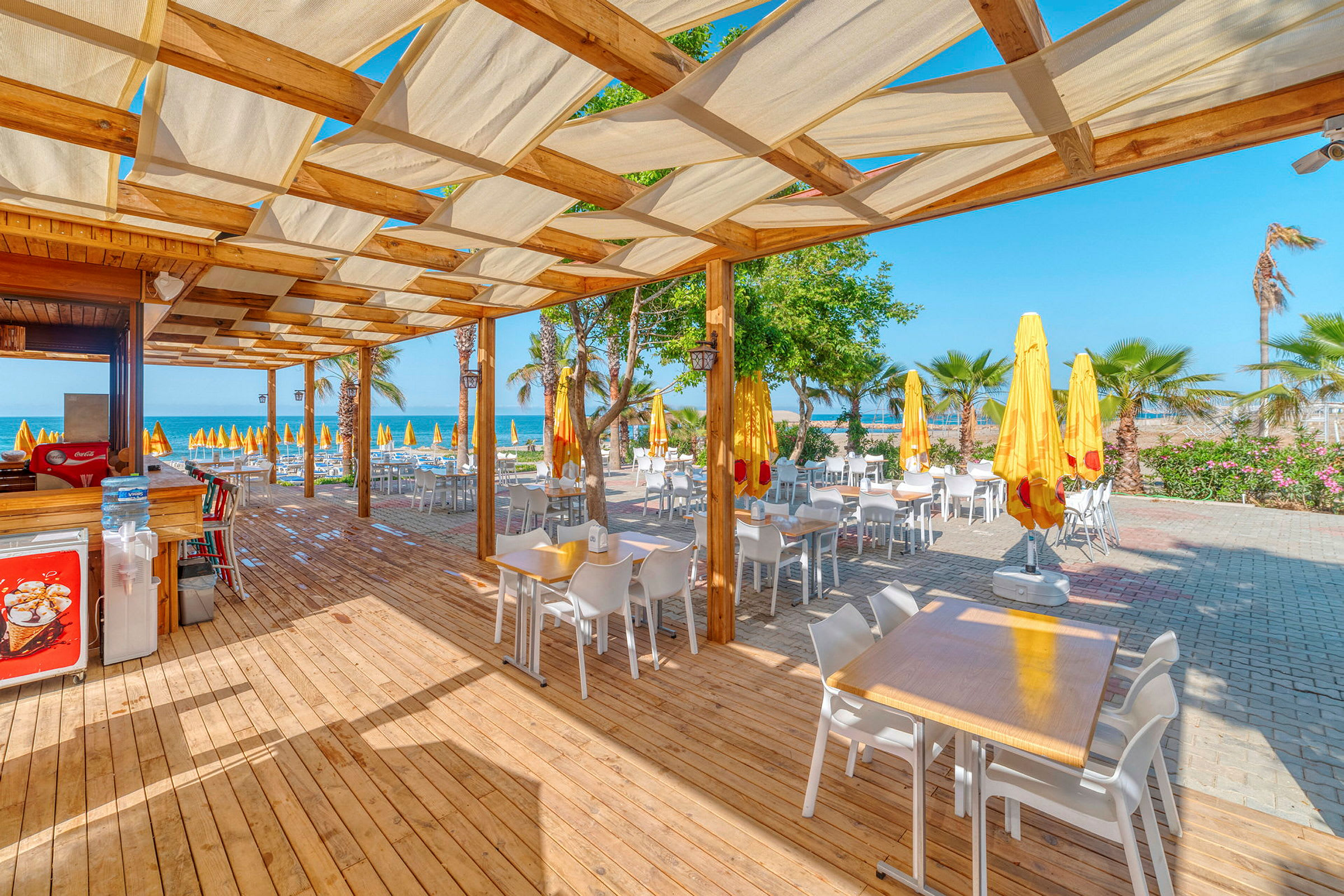 Udendørs spisested med borde og stole under en pergola ved stranden. (AI-genereret tekst)