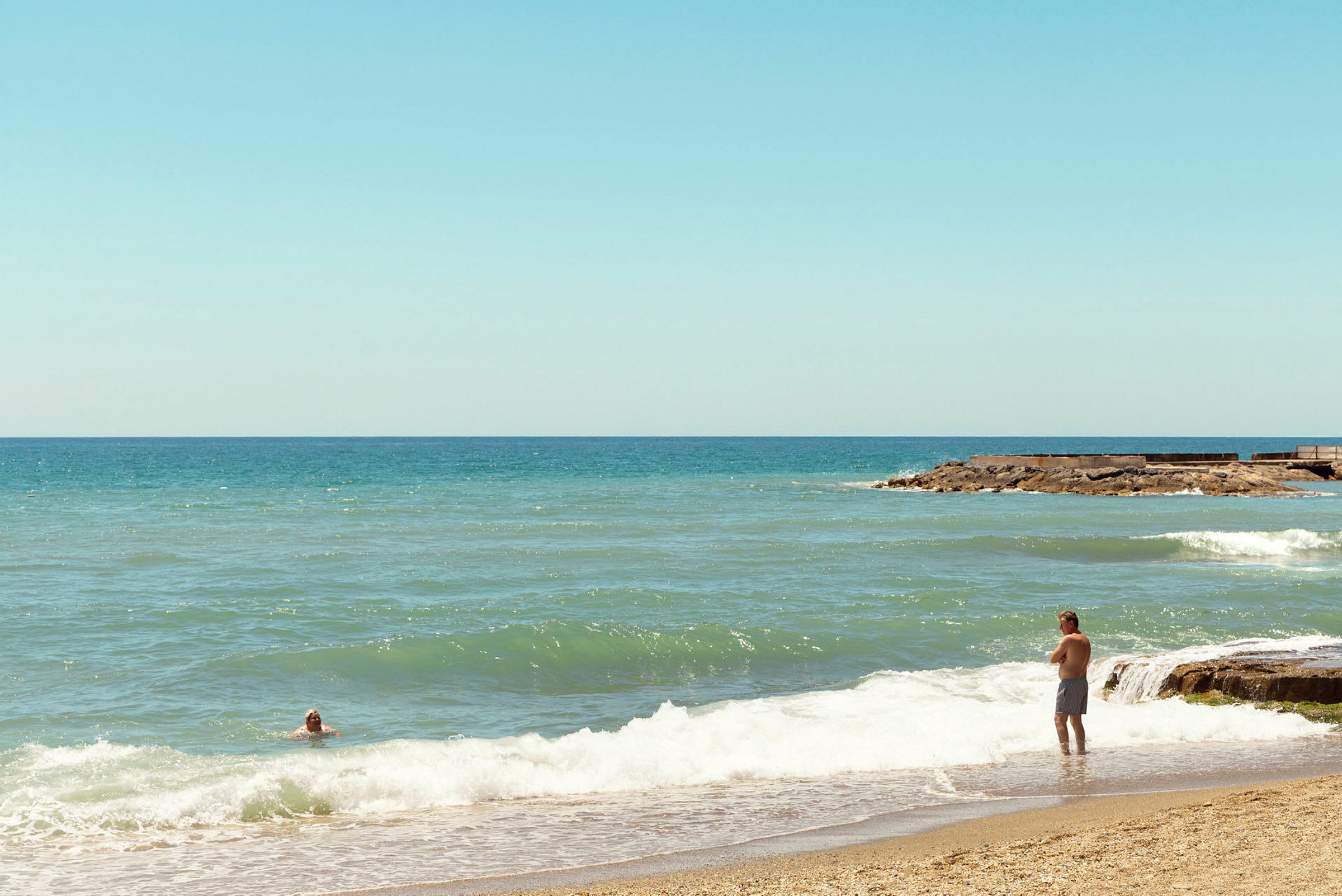 En strand med sand i forgrunden og bølger, der ruller ind. To personer er synlige på billedet, en står ved vandkanten og en svømmer i vandet. (AI-genereret tekst)