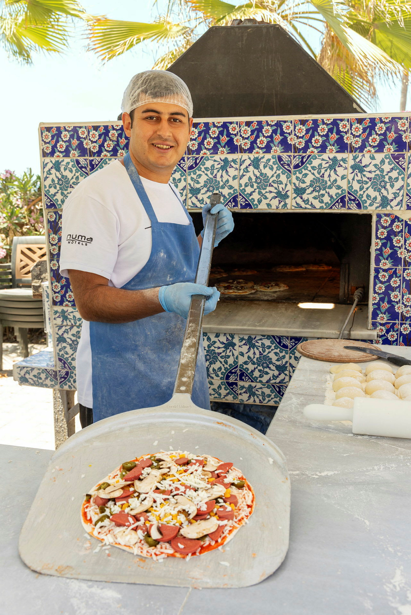En pizzabager holder en spade med en klargjort pizza foran en ovn dekoreret med blå og hvide fliser. (AI-genereret tekst)