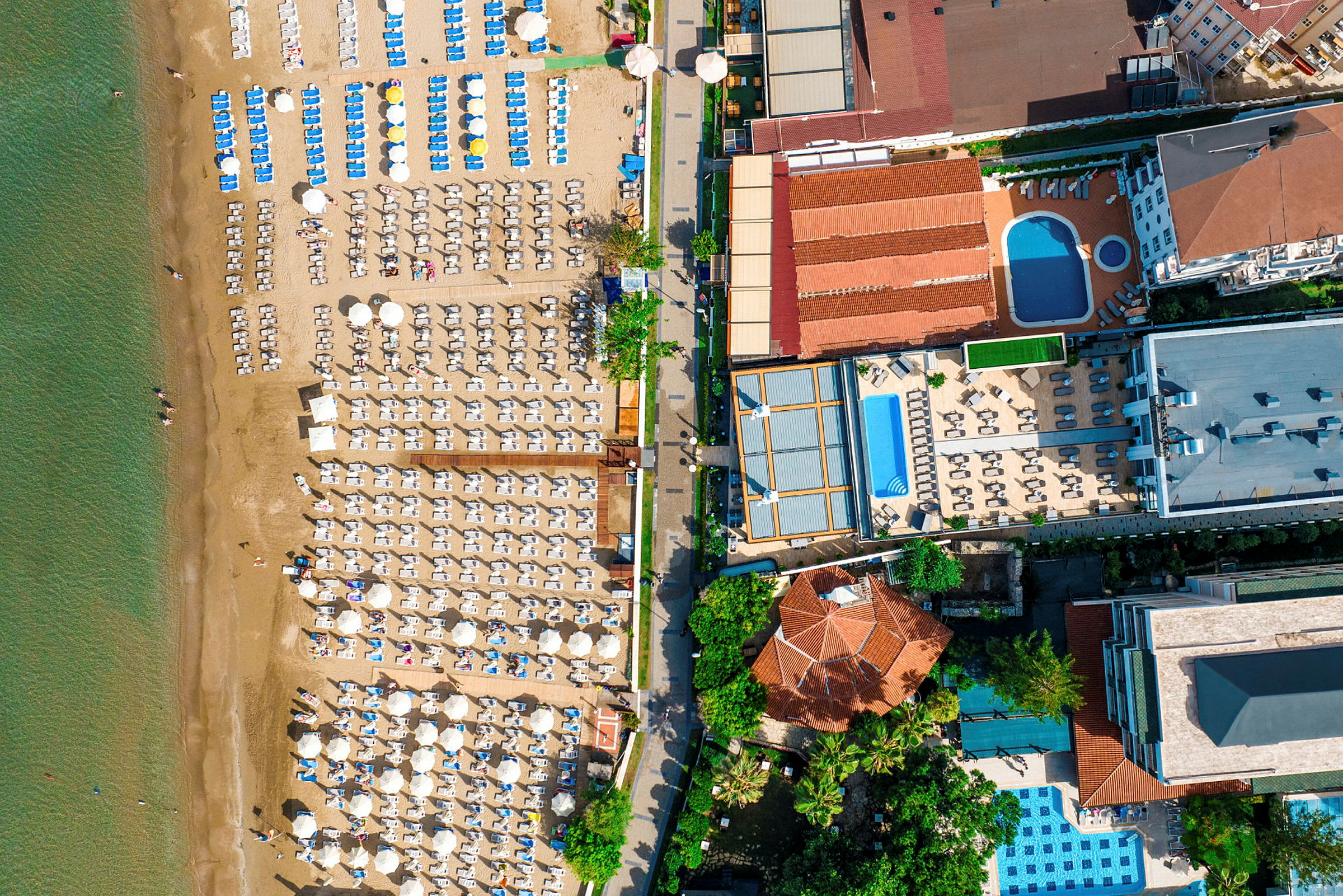 Luftfoto af en strand med liggestole, parasoller og tilstødende hotelområder med swimmingpools. (AI-genereret tekst)