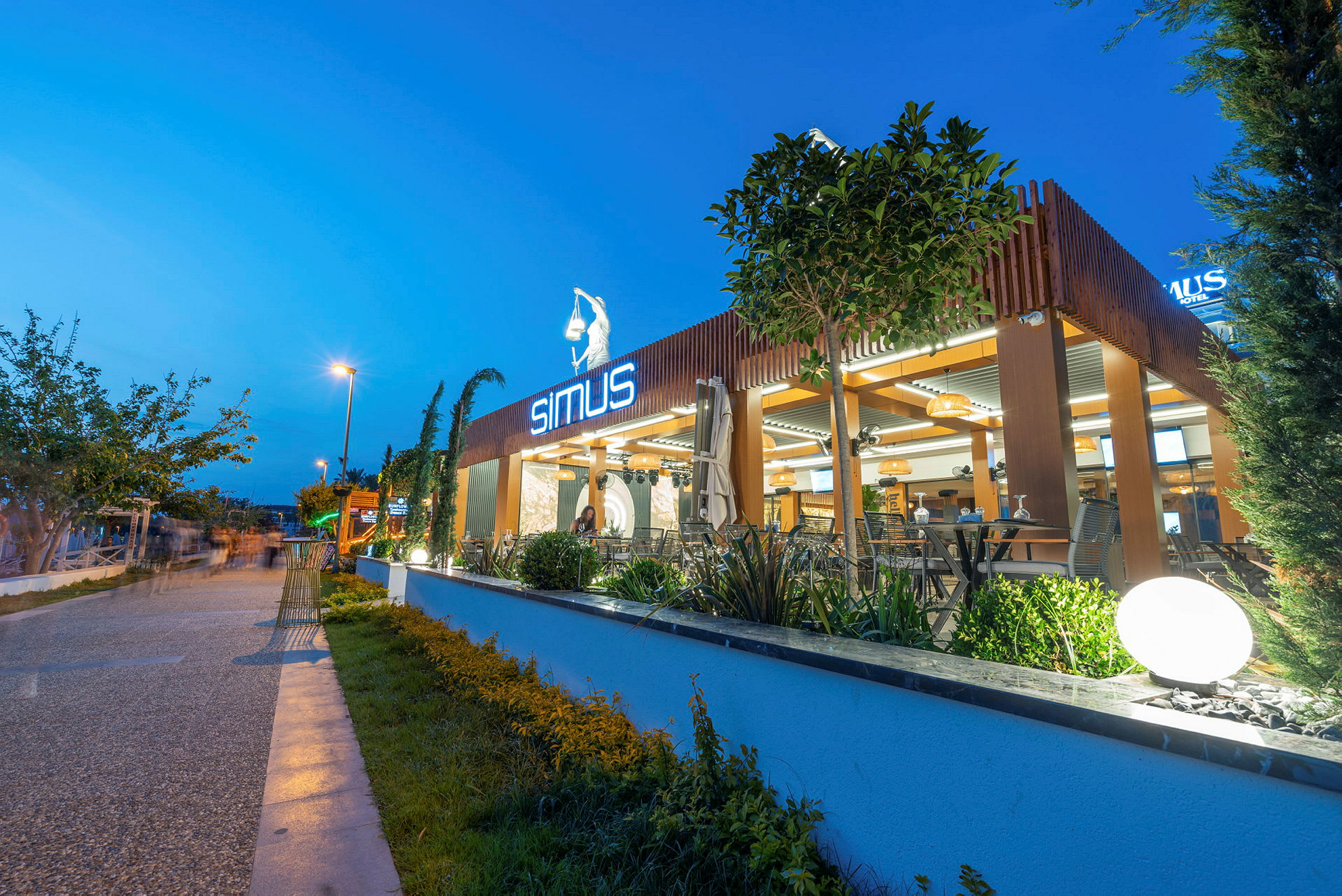 Moderne restaurant med neonskilt, udendørs område og planteudsmykning på en aftenoplyst gade. (AI-genereret tekst)