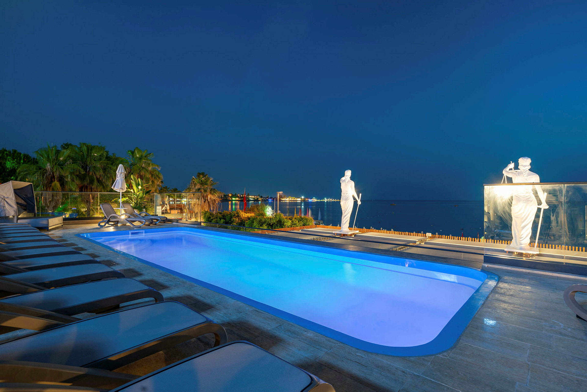 En oplyst pool omgivet af liggestole og palmer med statuer og havet i baggrunden. (AI-genereret tekst)