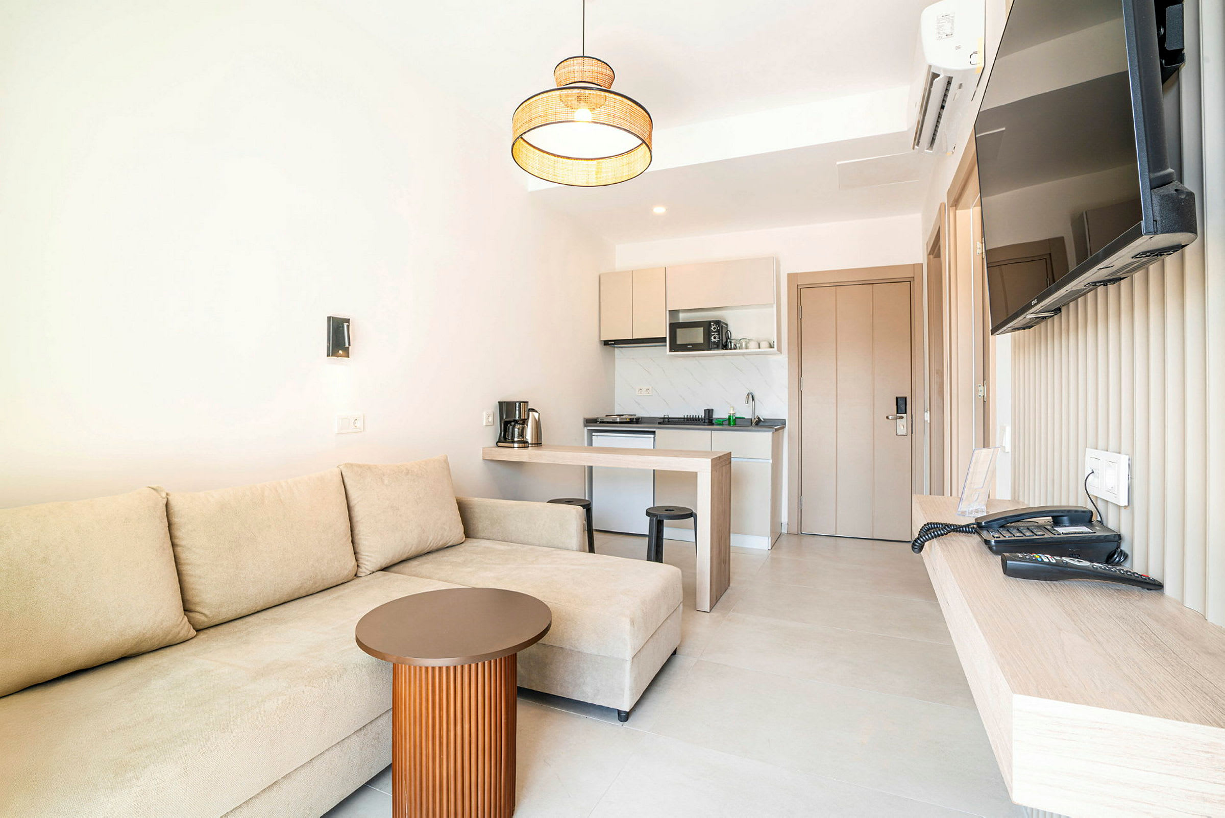 Et hotelværelse med en beige sofa, et lille bord, et køkken og et vægmonteret TV. (AI-genereret tekst)
