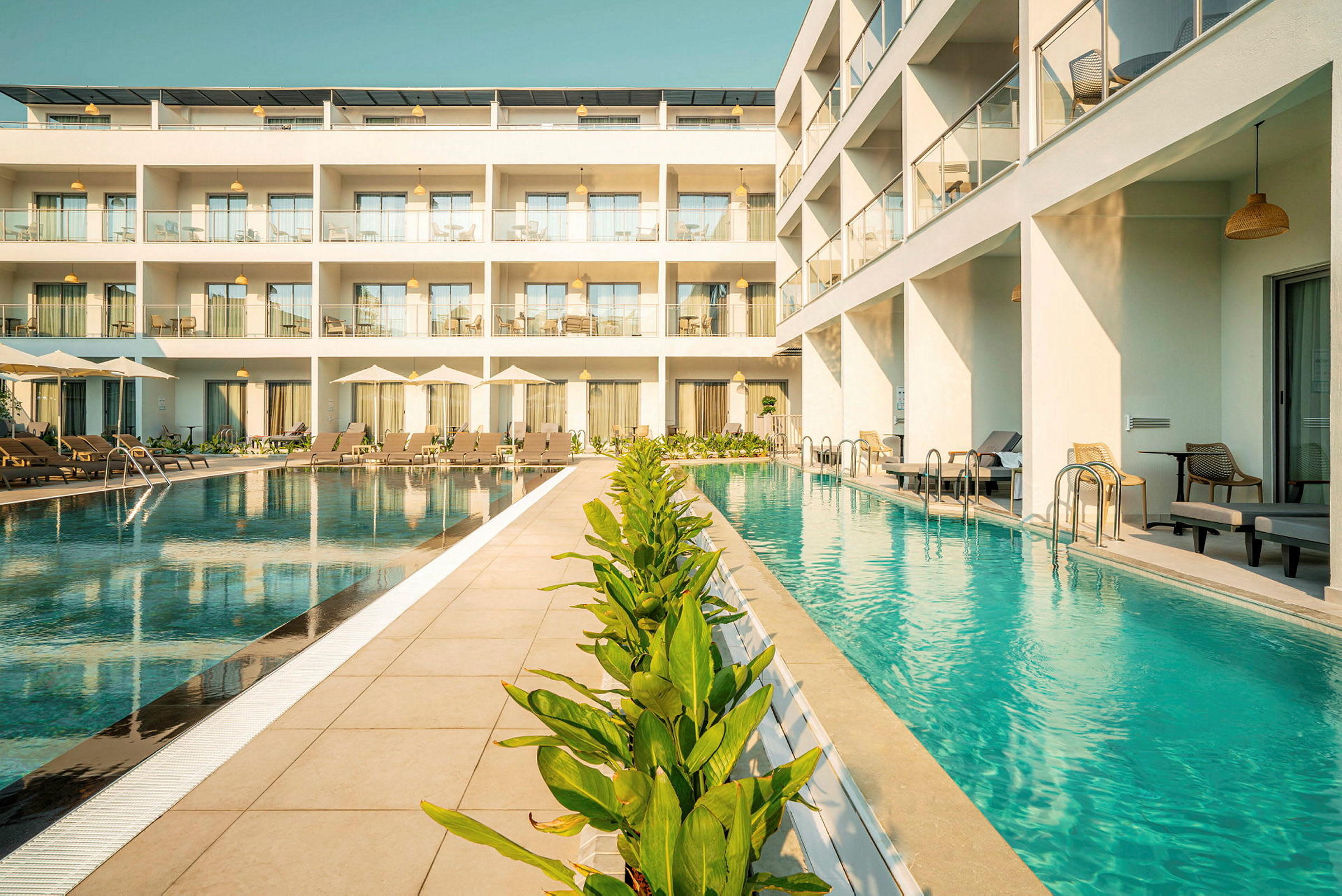 Billede af hotel med pool foran bygningen med liggestole og grønne planter. (AI-genereret tekst)