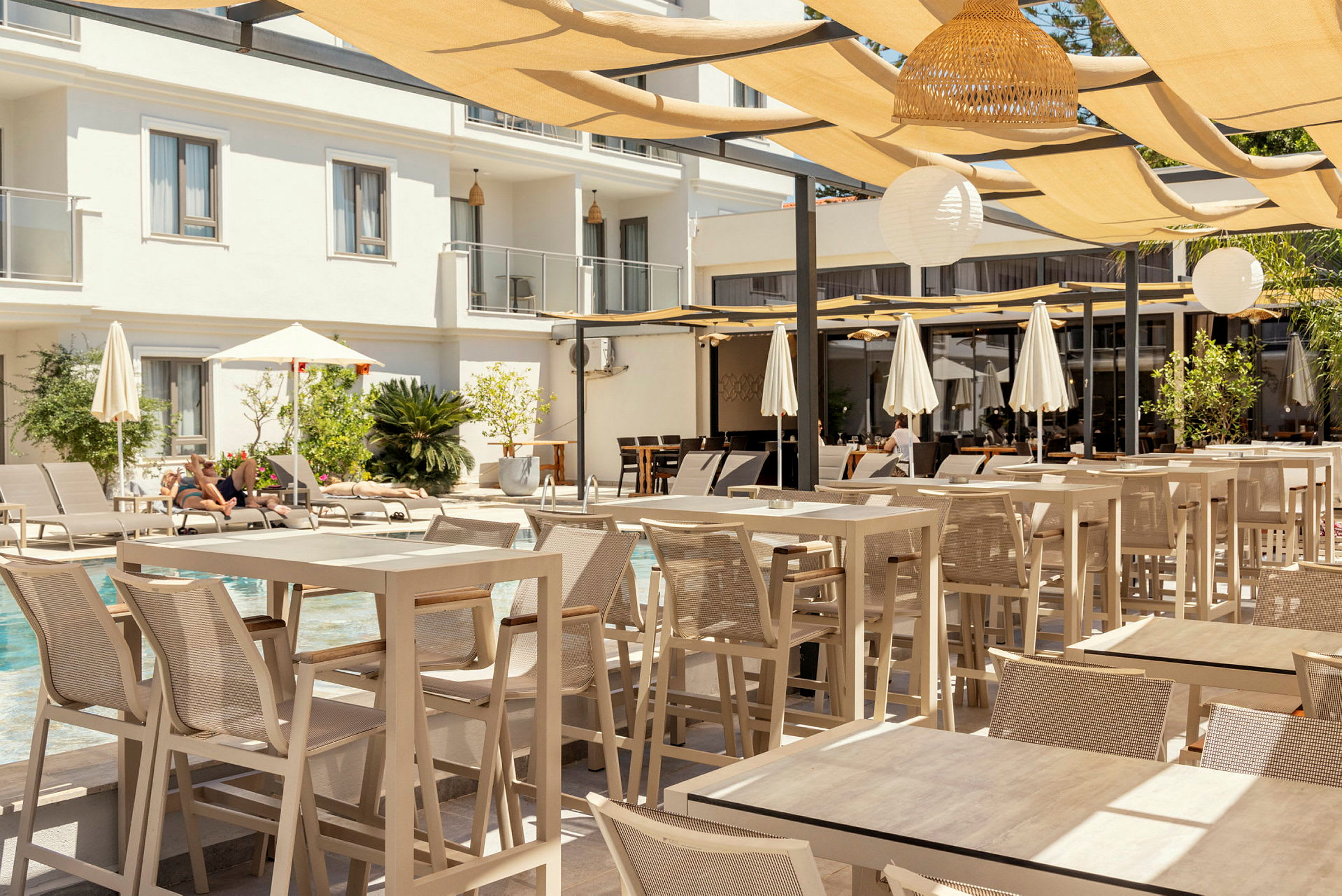 Et udendørsområde på et hotel med borde og stole under en skyggefuld pergola, med en pool og solsenge i baggrunden. (AI-genereret tekst)