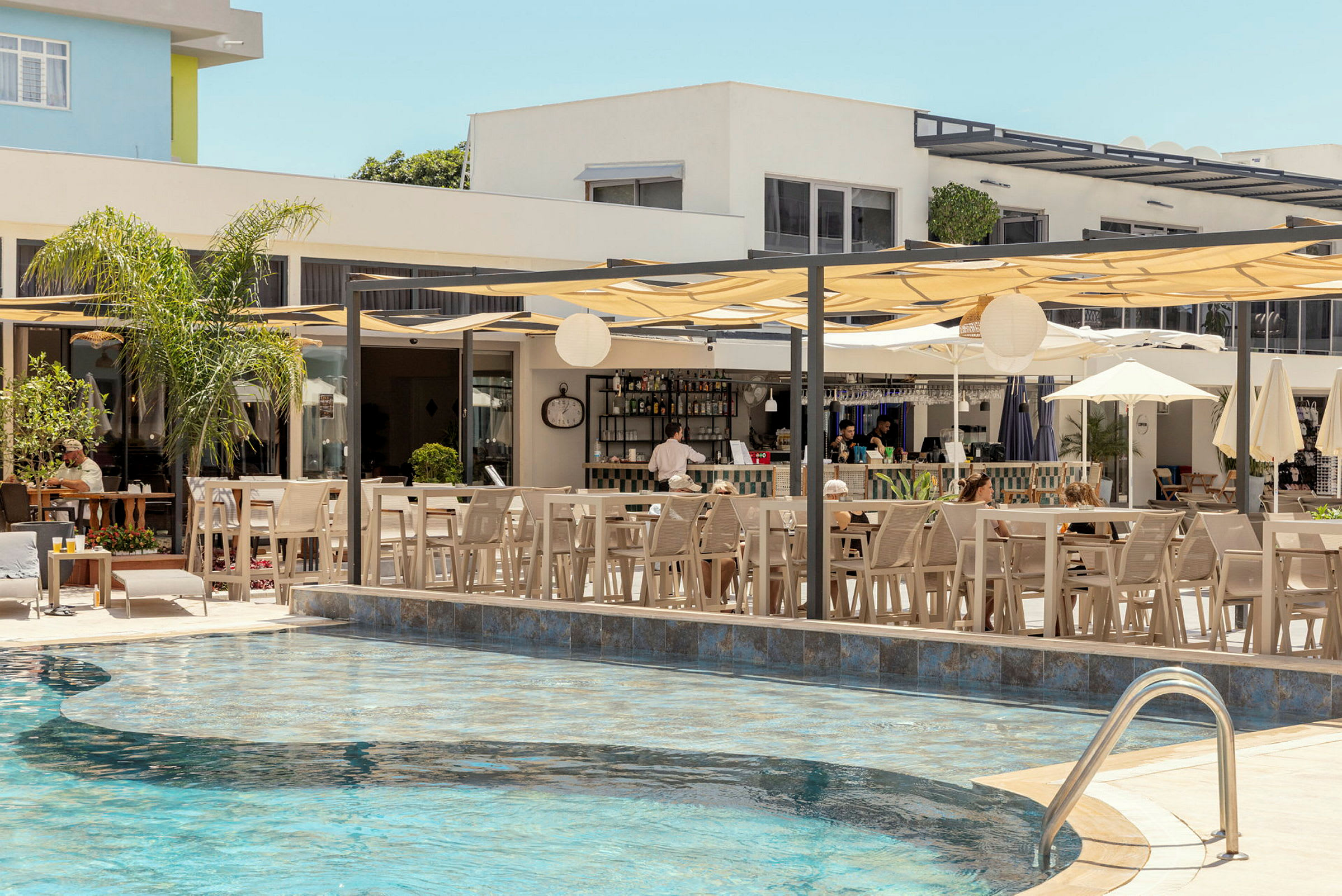 En udendørs swimmingpool med omkringliggende siddepladser og parasoller ved en bar i baggrunden. (AI-genereret tekst)