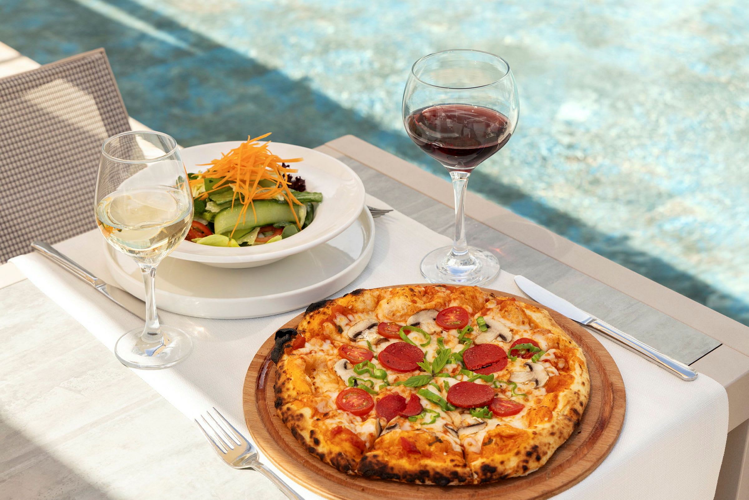 En tallerken med salat og et glas hvidvin ved siden af en pizza med pålæg og et glas rødvin, på et bord med baggrund af en pool. (AI-genereret tekst)