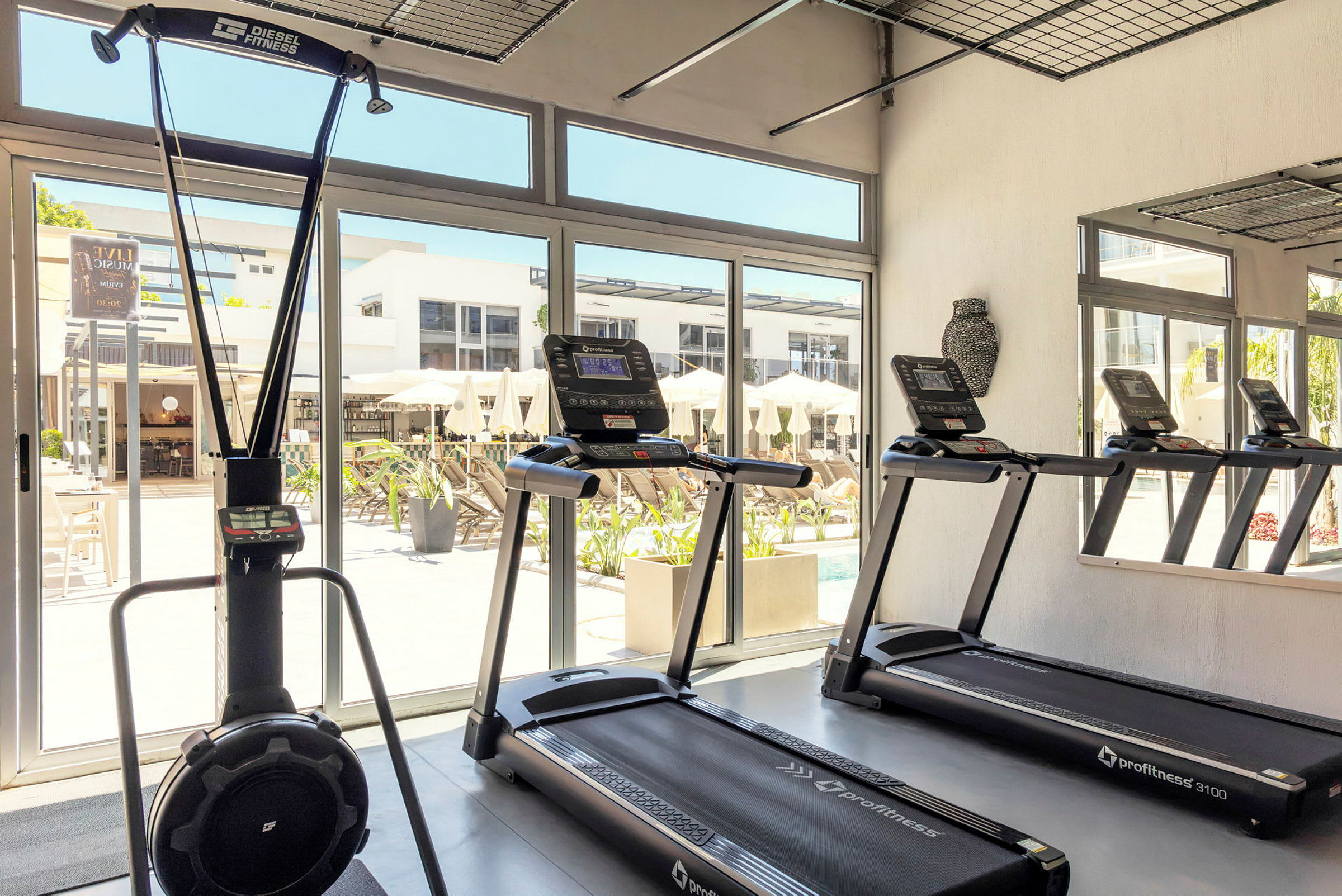Et fitnessrum med træningsmaskiner, herunder løbebånd og en stakmaskine. Store vinduer giver udsigt over en terrasse med parasoller. (AI-genereret tekst)