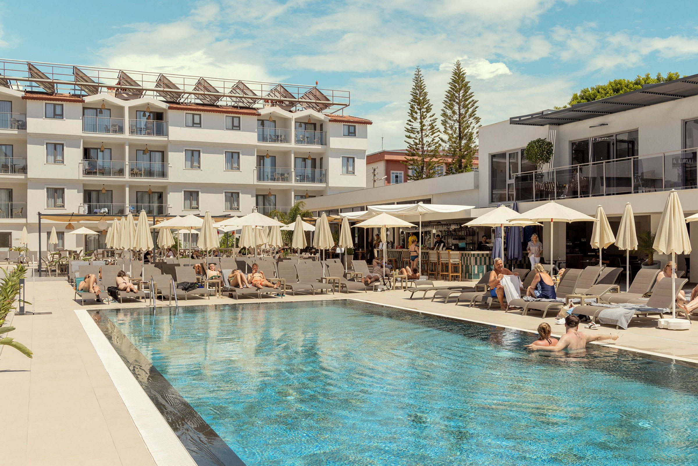 En hotelpool med liggestole og parasoller, omgivet af en hvid bygning og barer. (AI-genereret tekst)