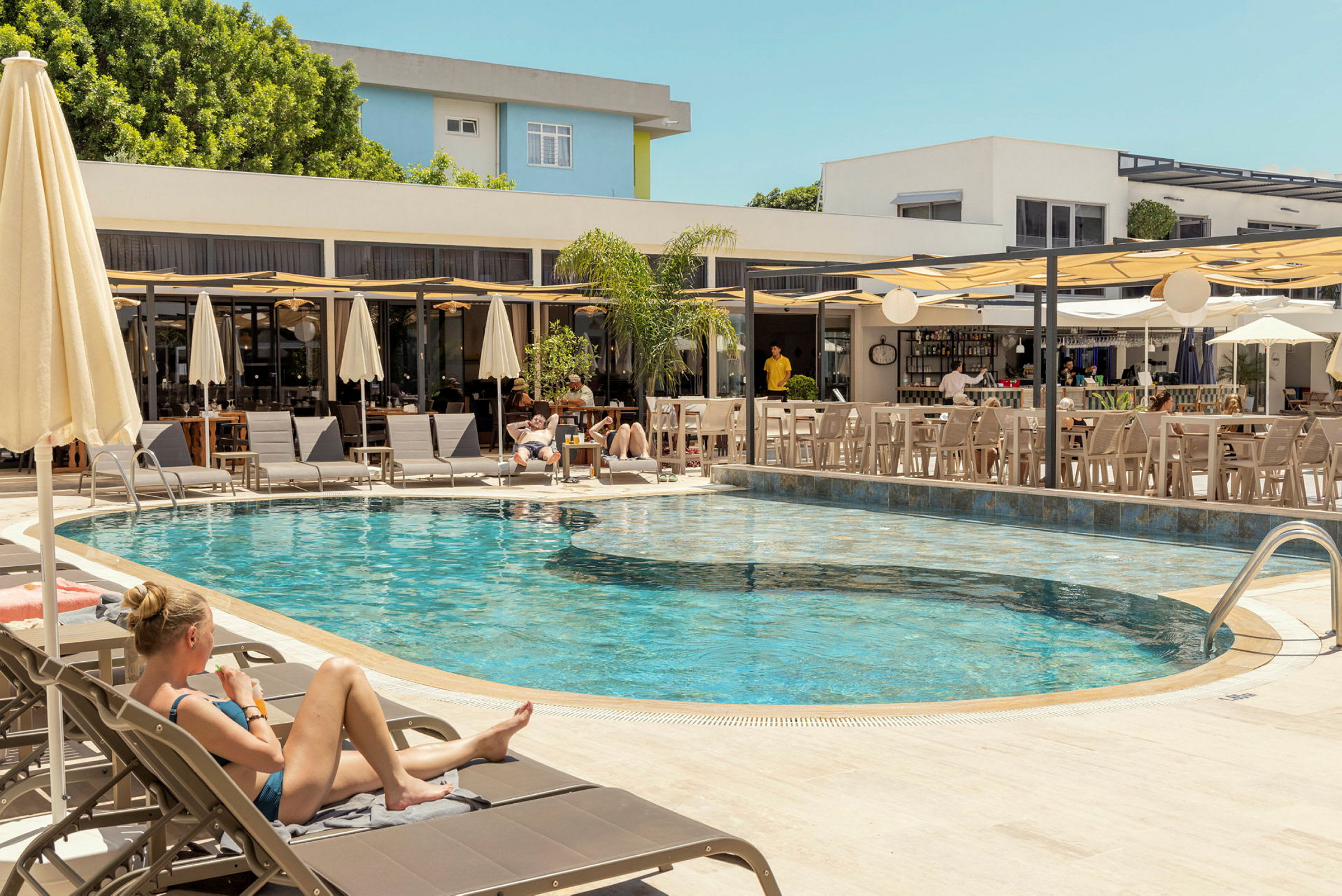 En udendørs pool omgivet af liggestole og parasoller, med en tilstødende bar og siddepladser. (AI-genereret tekst)