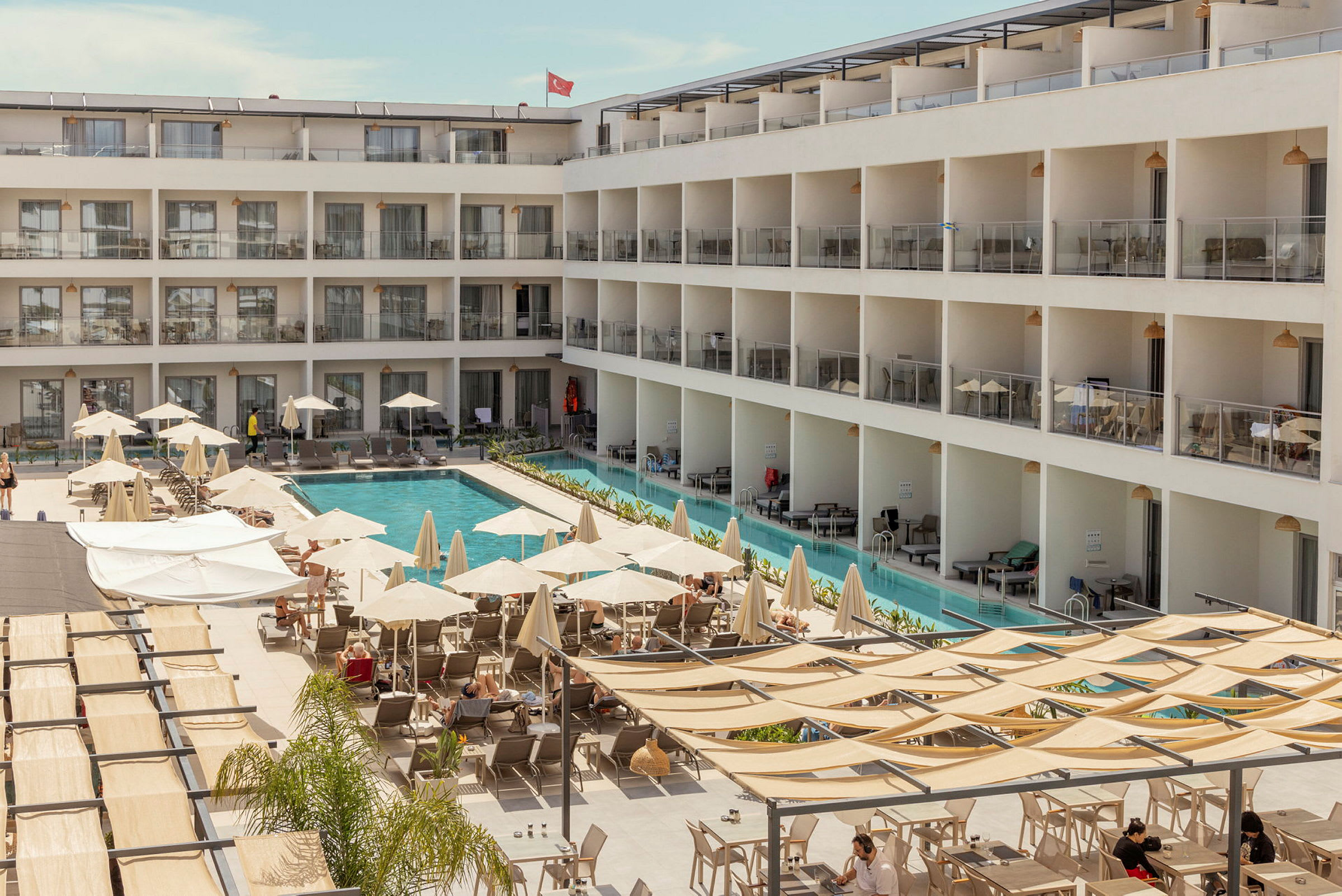 En hotelbygning med en pool i midten samt liggestole og parasoller omkring. (AI-genereret tekst)