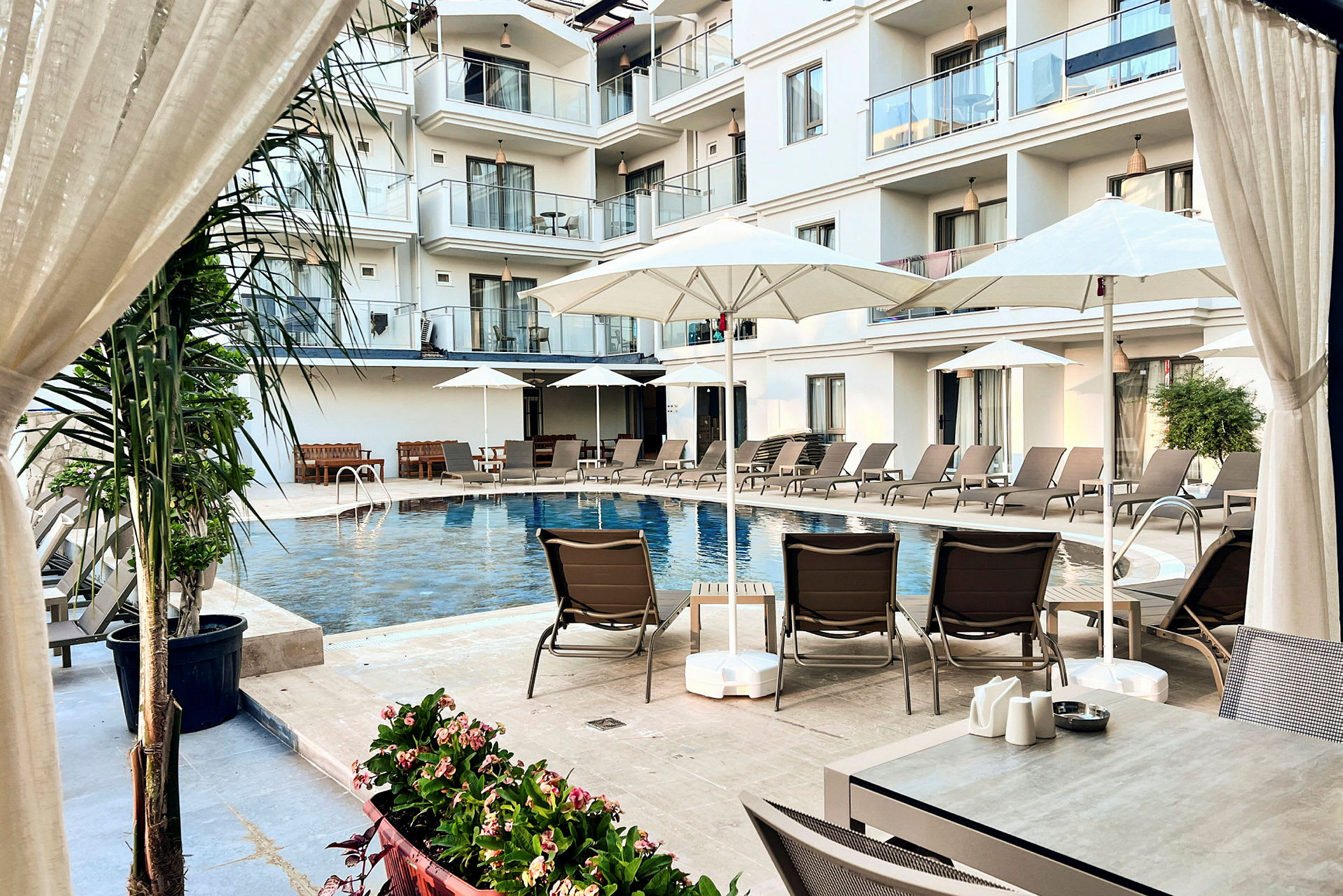 Billedet viser en swimmingpool ved et hotel omgivet af liggestole, parasoller og altaner i baggrunden. (AI-genereret tekst)