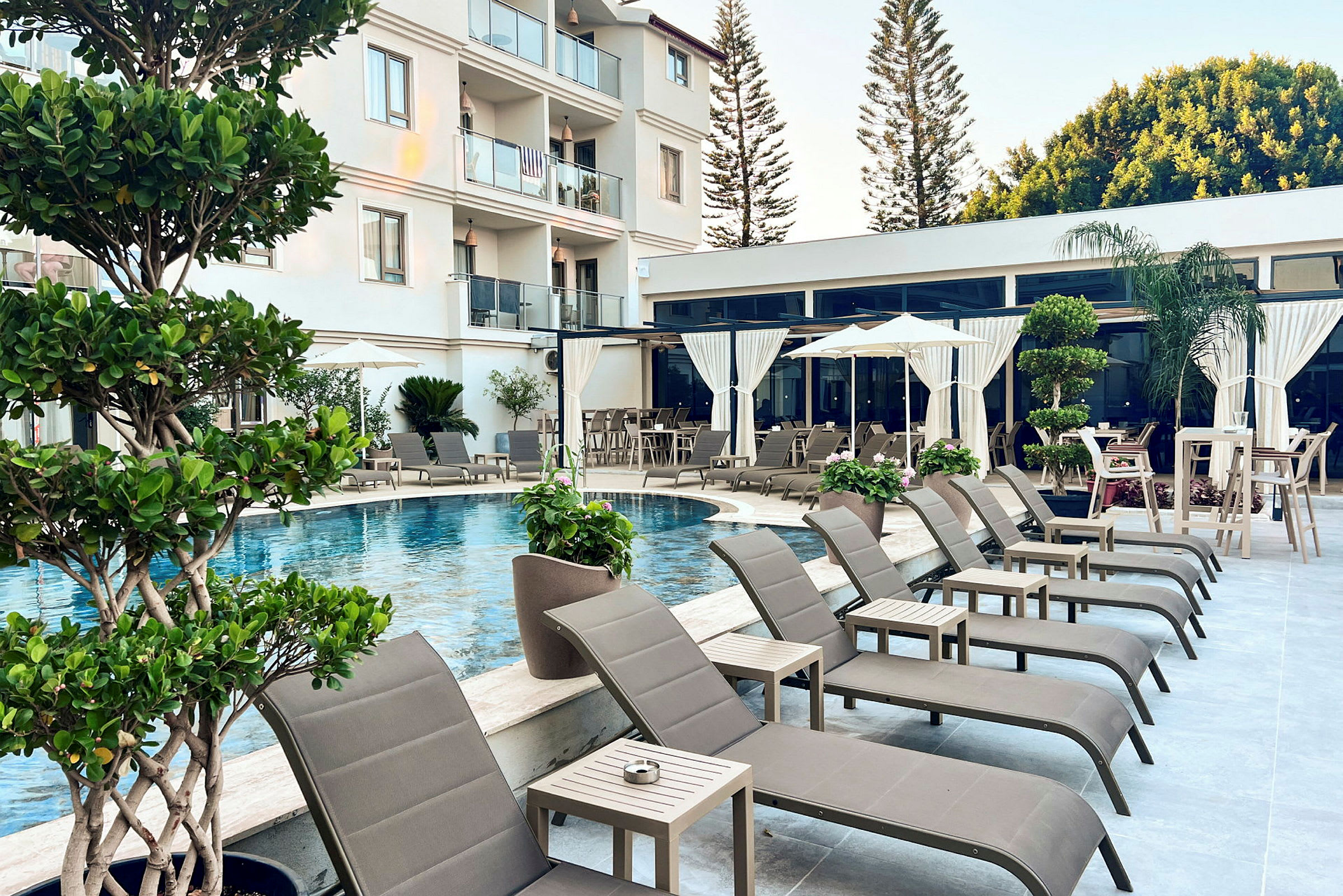 Poolområde med liggestole, parasoller og borde samt omgivende planter ved et hotel. (AI-genereret tekst)