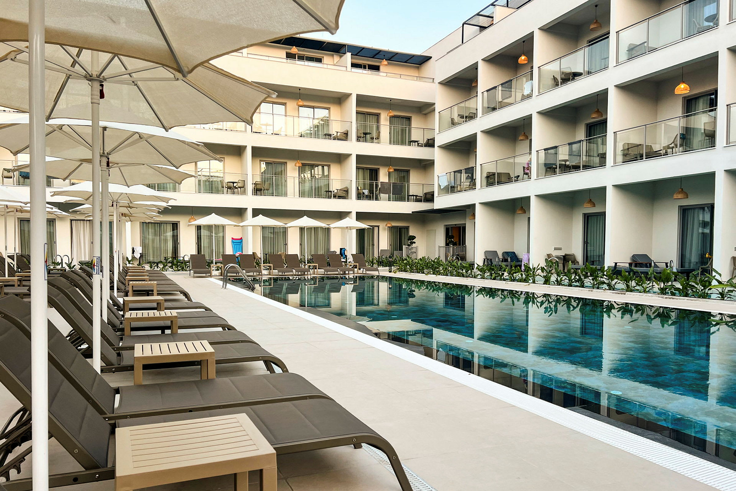 Et hotel med en pool omgivet af liggestole og parasoller samt altaner ovenover. (AI-genereret tekst)
